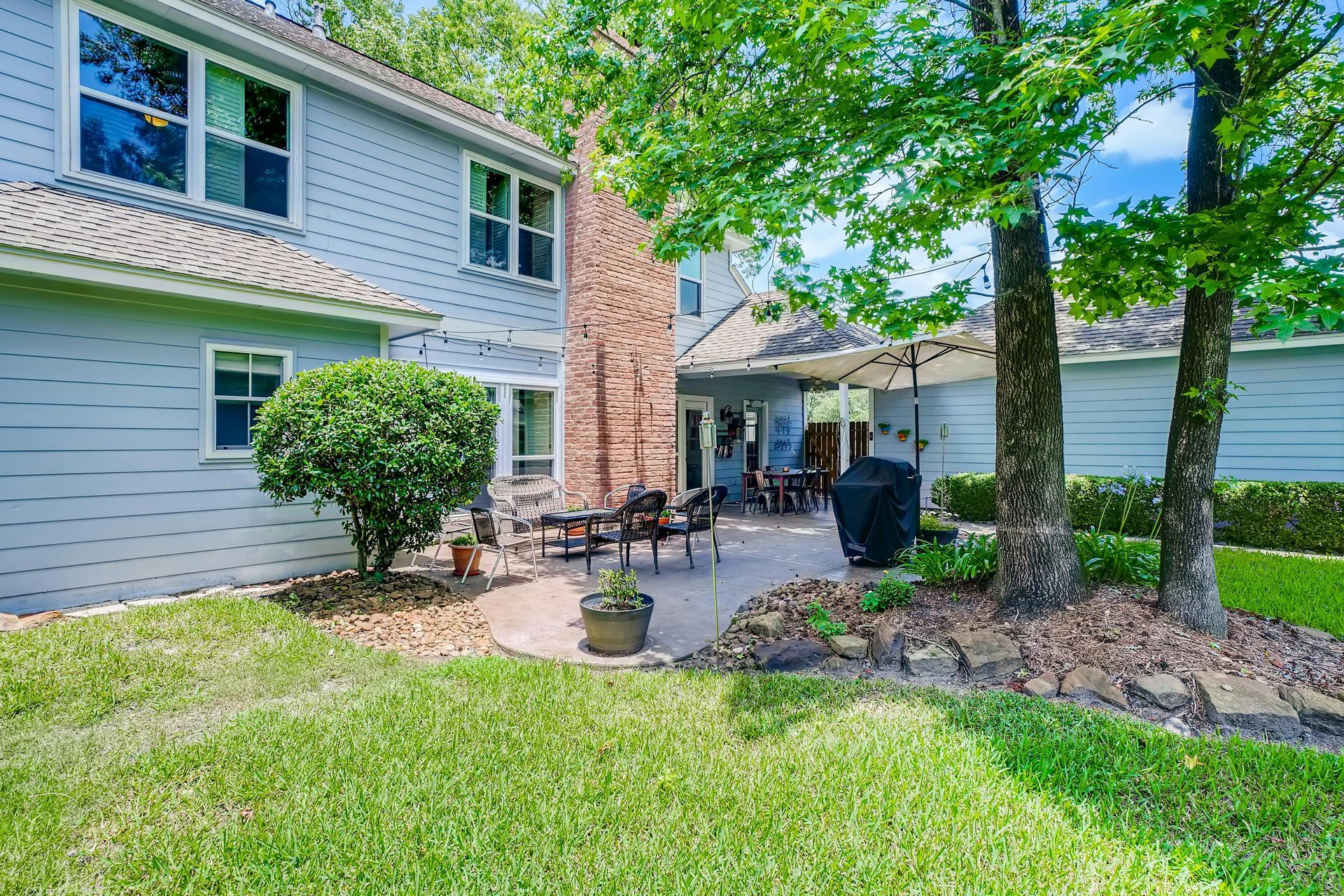 Backyard with patio, trees, and house. Green grass in foreground, blue siding on the house.