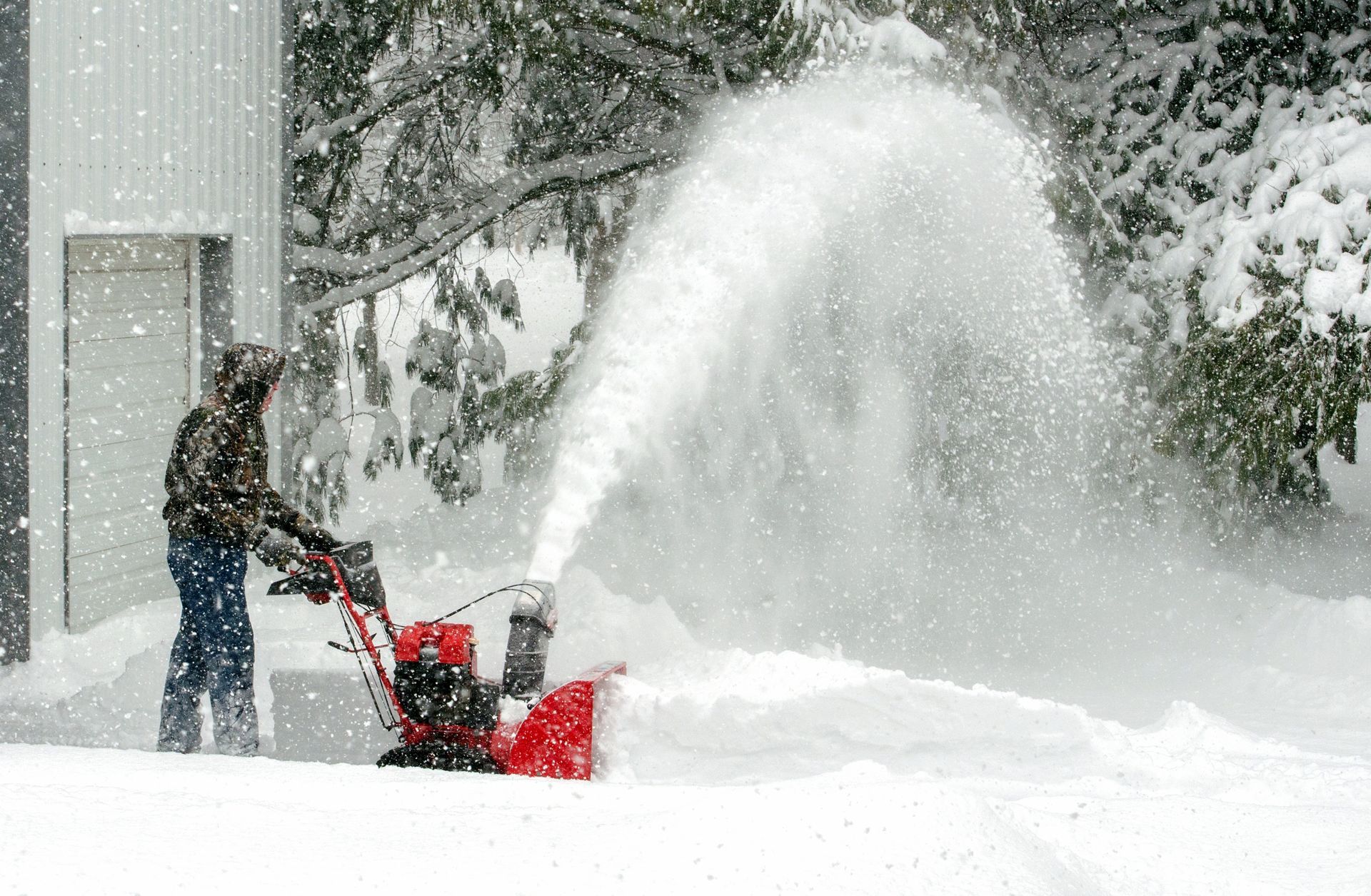 Person using a snowblower in a snowy yard, white snow being ejected into the air.