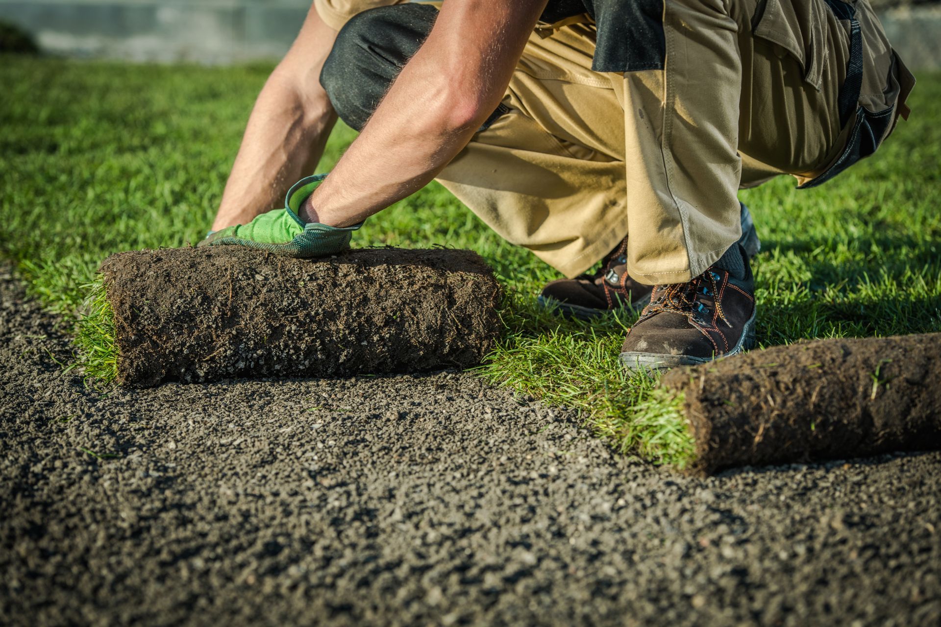 Person installing sod rolls on a gravel path, wearing gloves and tan pants.