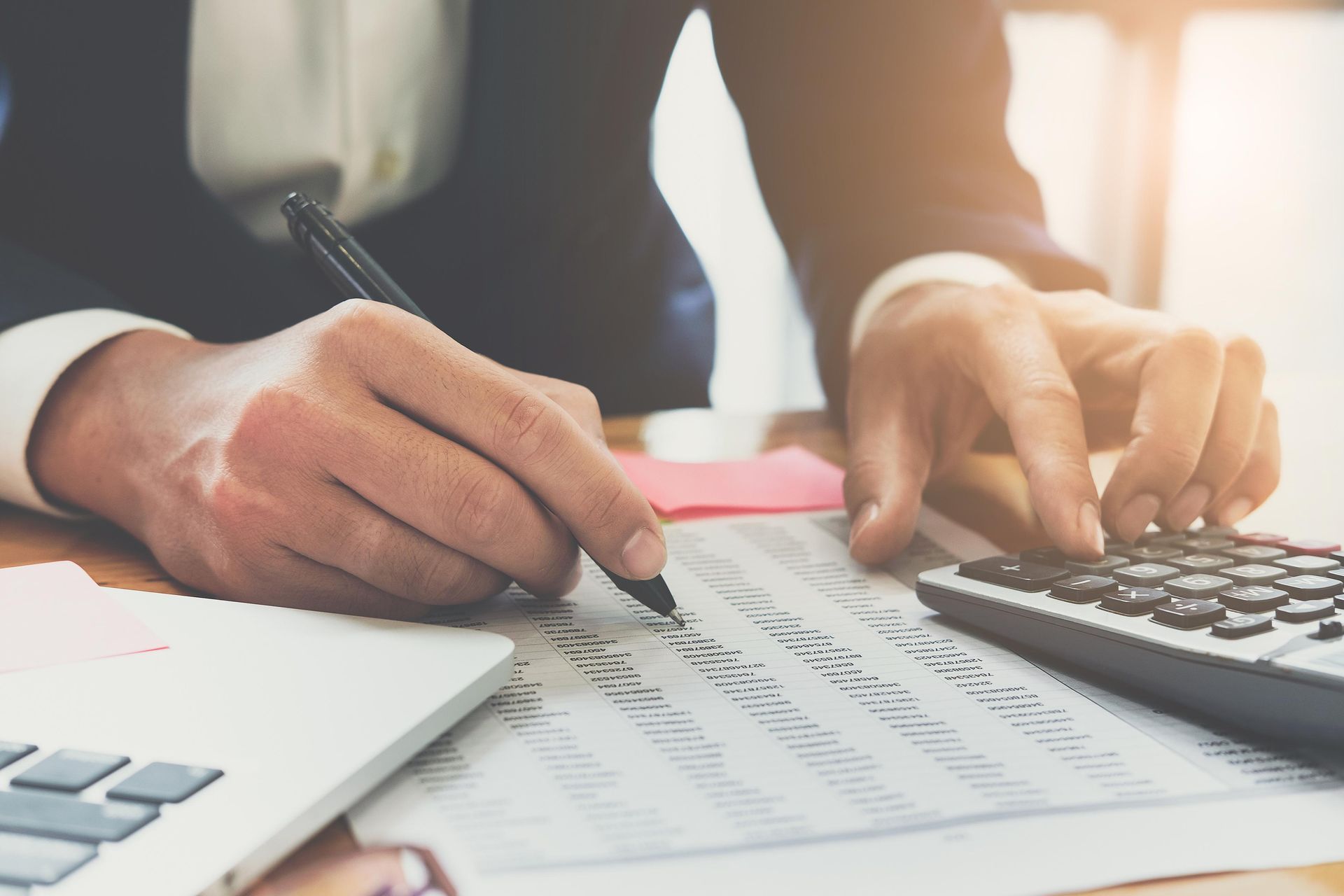 Person using a calculator and pen to work on financial documents at a desk with a laptop.