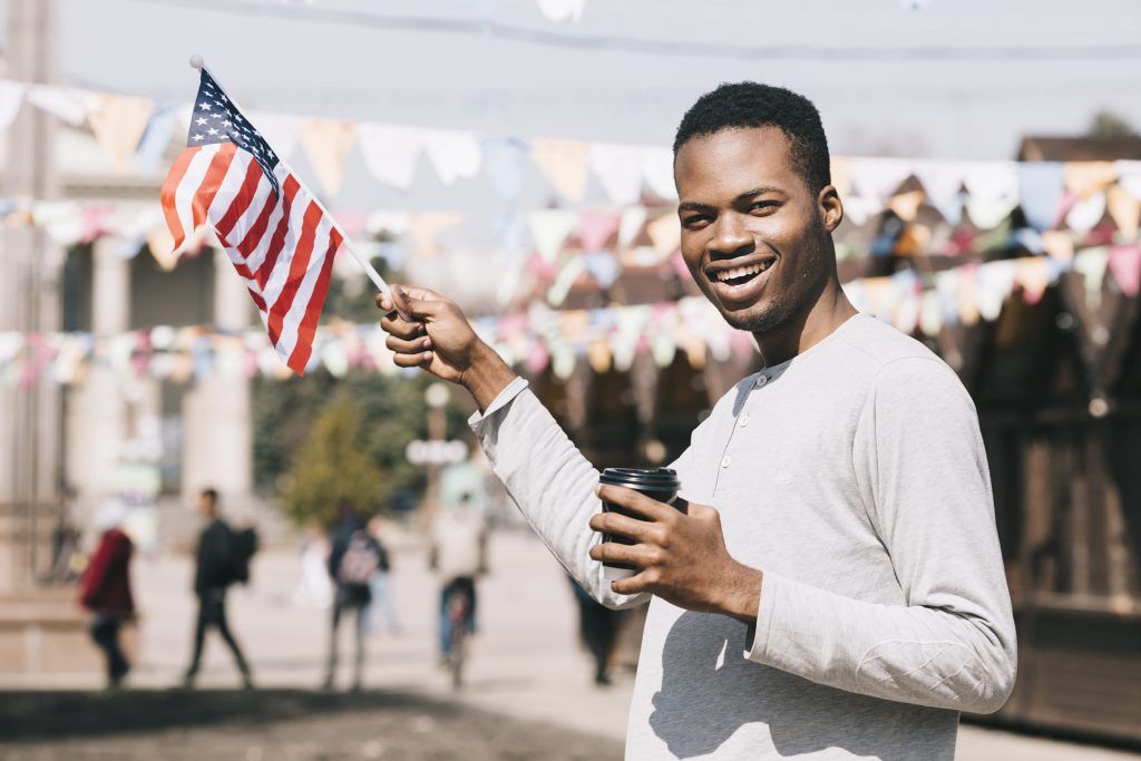 A smiling person holds a US flag and a coffee cup, standing outdoors under festive bunting.