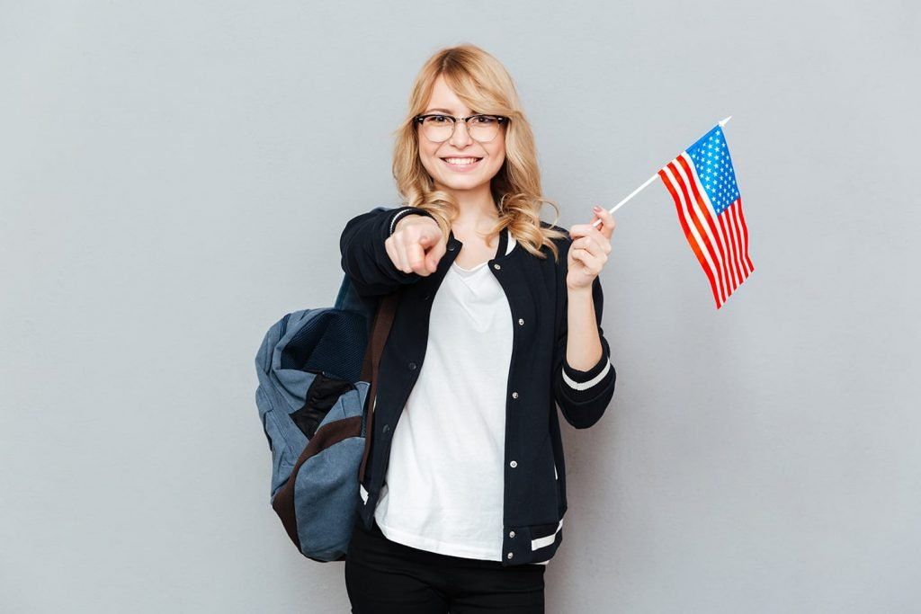 A student with a backpack smiles and points forward while holding a small American flag against a grey background.