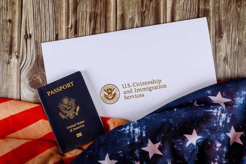 A U.S. passport and USCIS document envelope rest on a wooden surface next to an American flag.