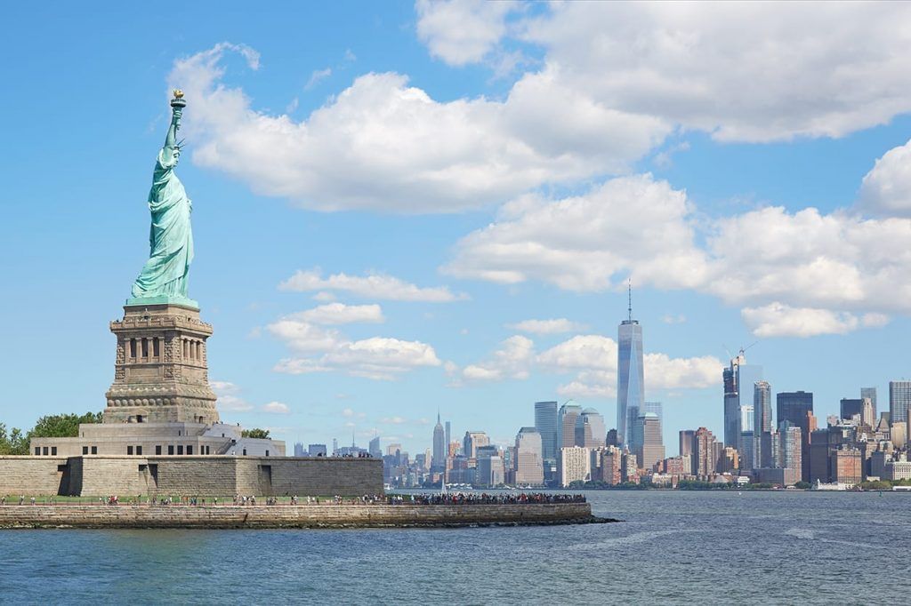 The Statue of Liberty standing on its pedestal, with the New York City skyline visible in the background under a blue sky.