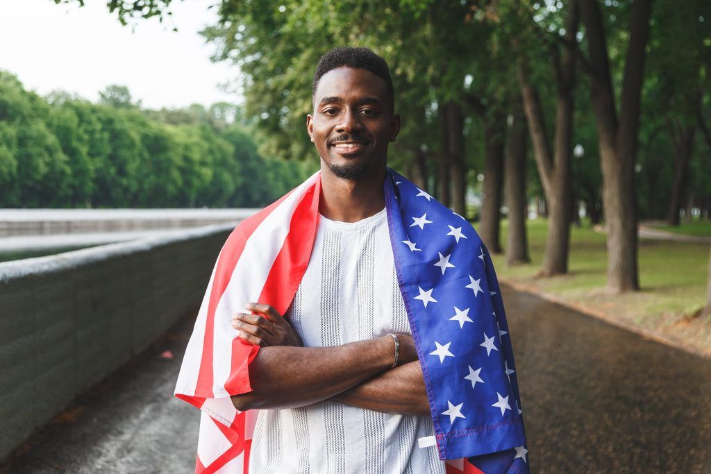 A man smiles with his arms crossed, wearing an American flag draped over his shoulders outdoors on a path lined with trees.