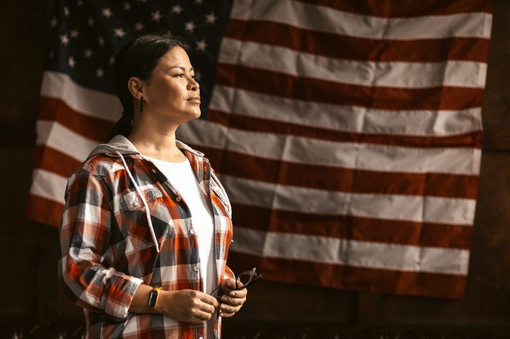 A person in a plaid shirt stands looking to the side against the backdrop of a large American flag.