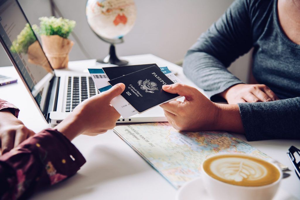 Two people hold a passport and boarding pass over a map, with a laptop, globe, and coffee cup on a desk.