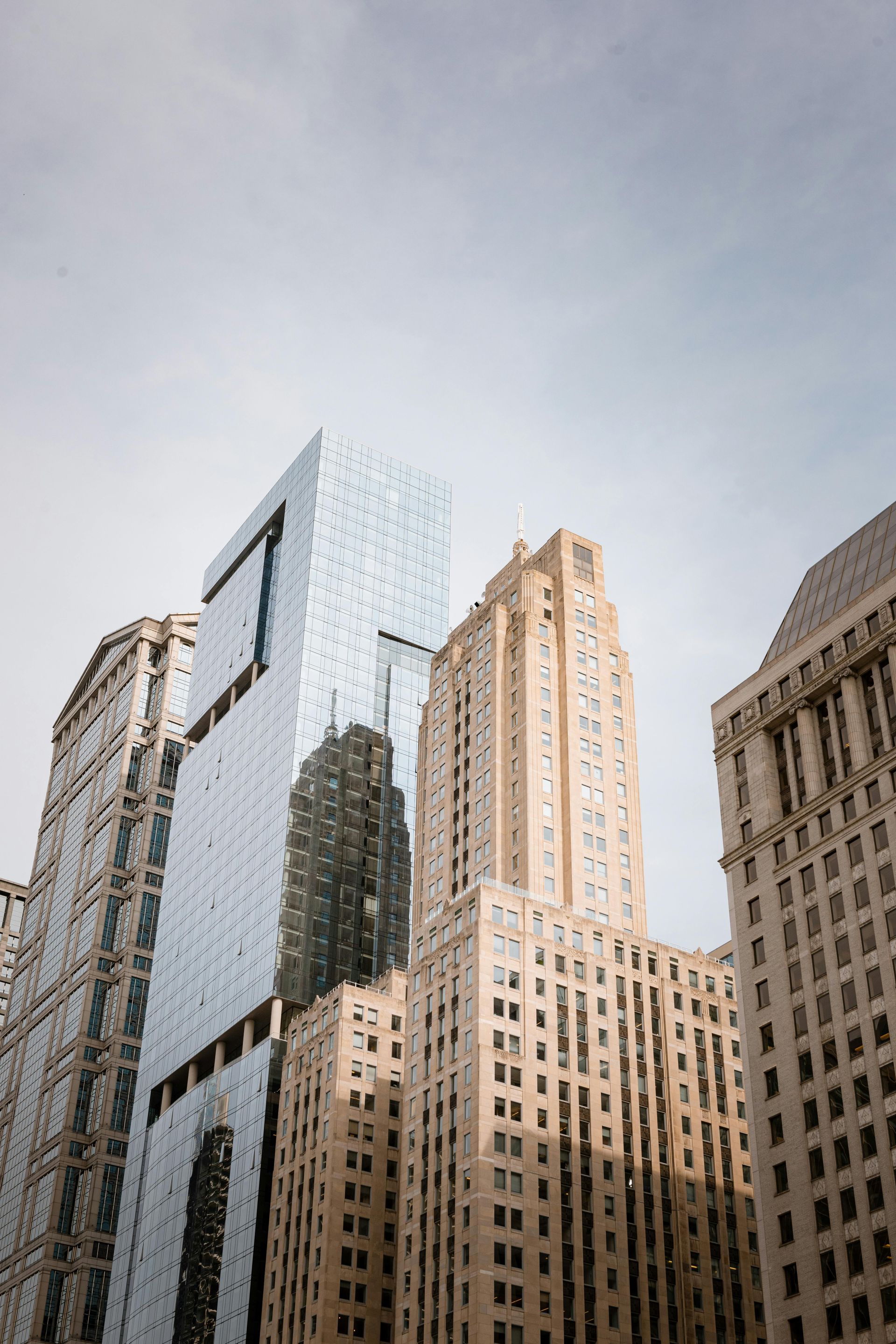 Low-angle view of four tall, architectural skyscrapers of varying modern and classic styles against a pale, overcast sky.