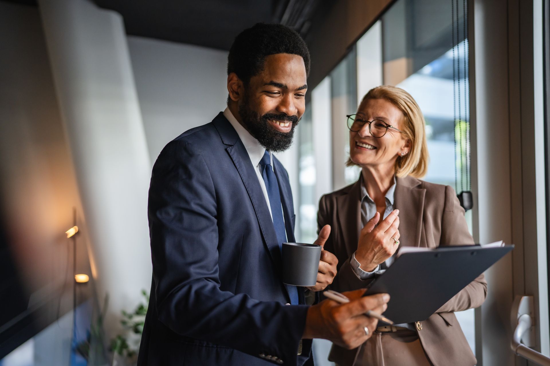 Two professionals in formal attire discussing work and holding a document in a bright, modern office hallway.
