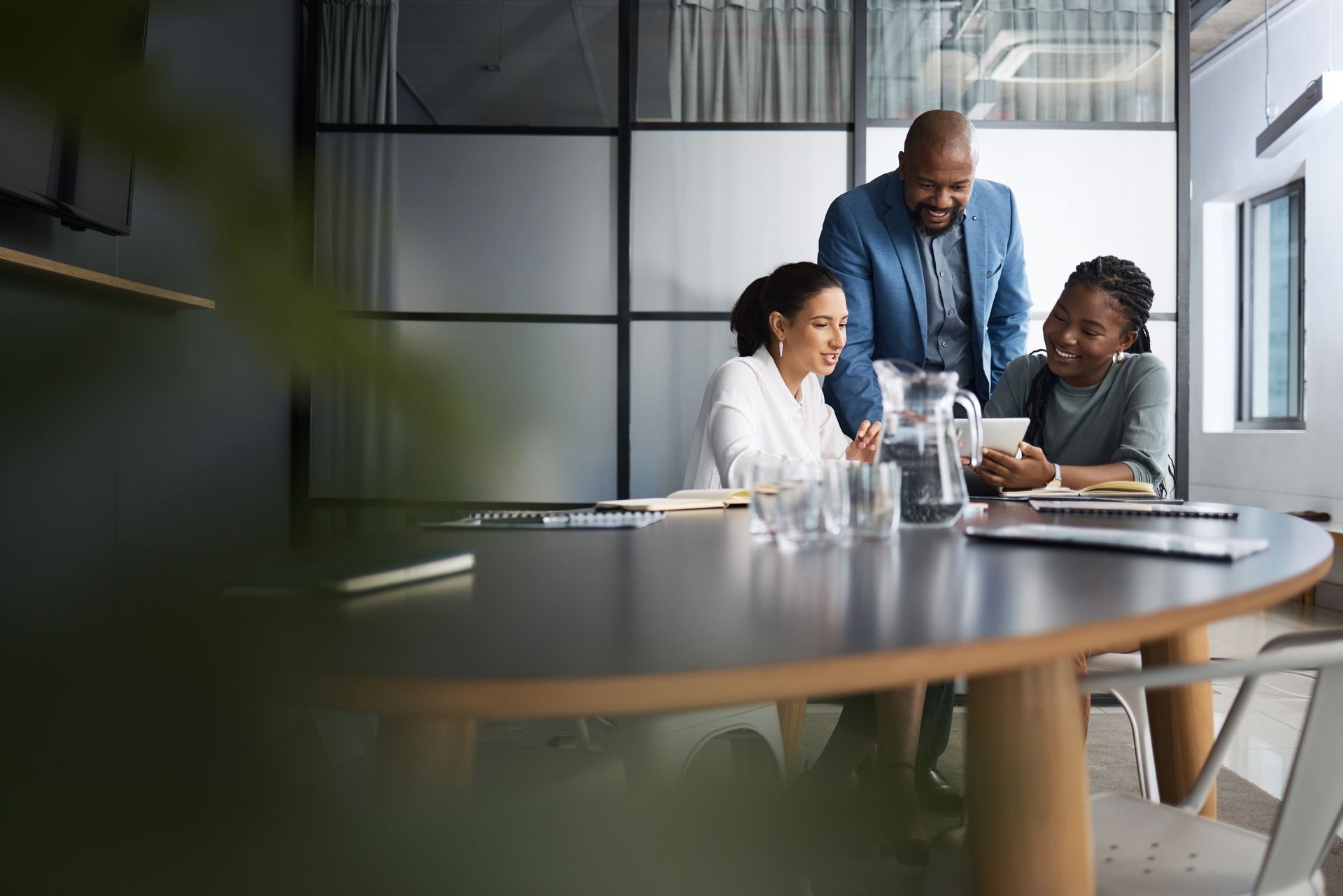 Three professionals collaborate around a wooden table in a bright, modern office with glass walls.