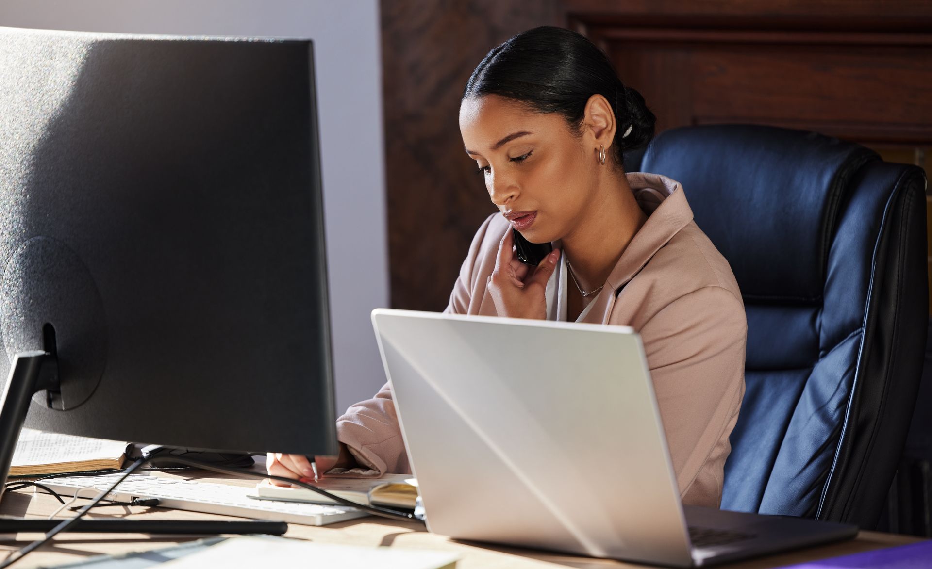 A focused professional wearing a pink blazer speaks on a phone while working at a desk with a computer and laptop.