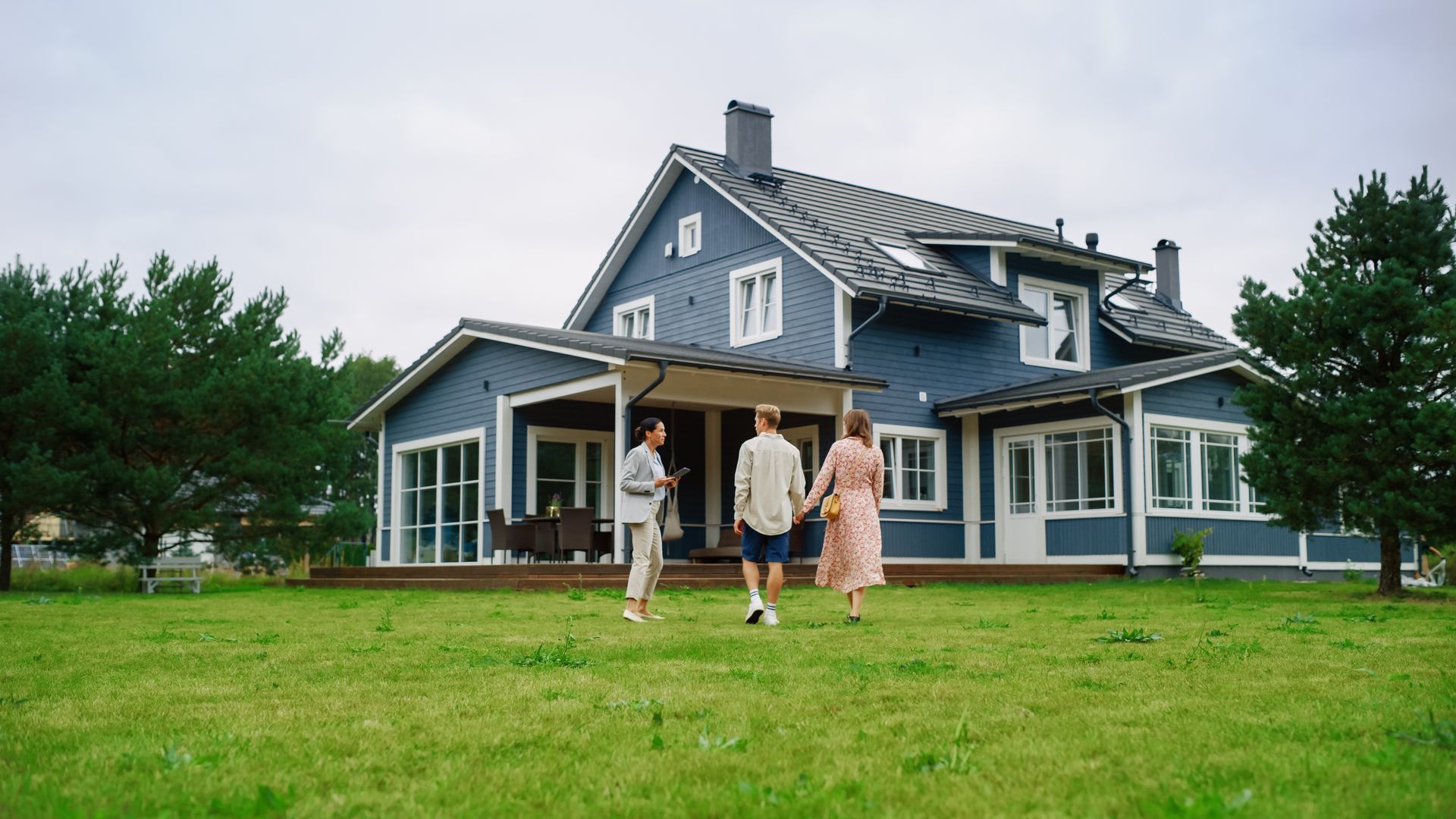 Three people walk across a grassy lawn toward a blue two-story house with a patio under a cloudy sky.