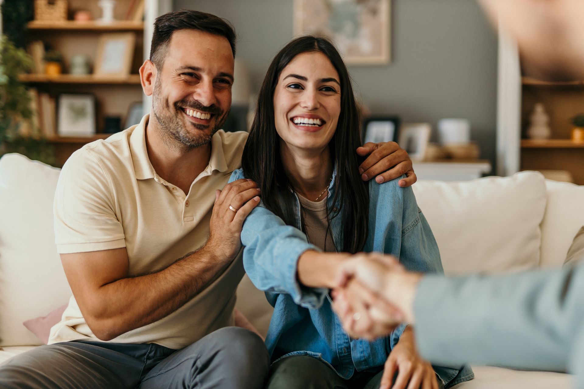 A couple smiles while sitting on a couch, shaking hands with an unseen person in a brightly lit, cozy living room.