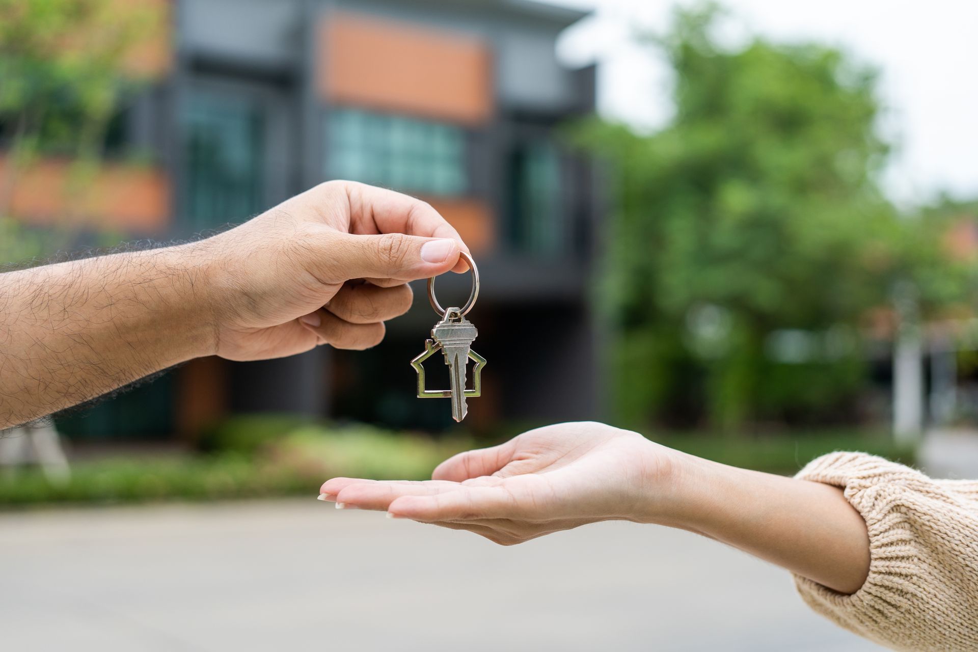 A hand hands a key with a house-shaped keychain to another person in front of a blurred residential building.