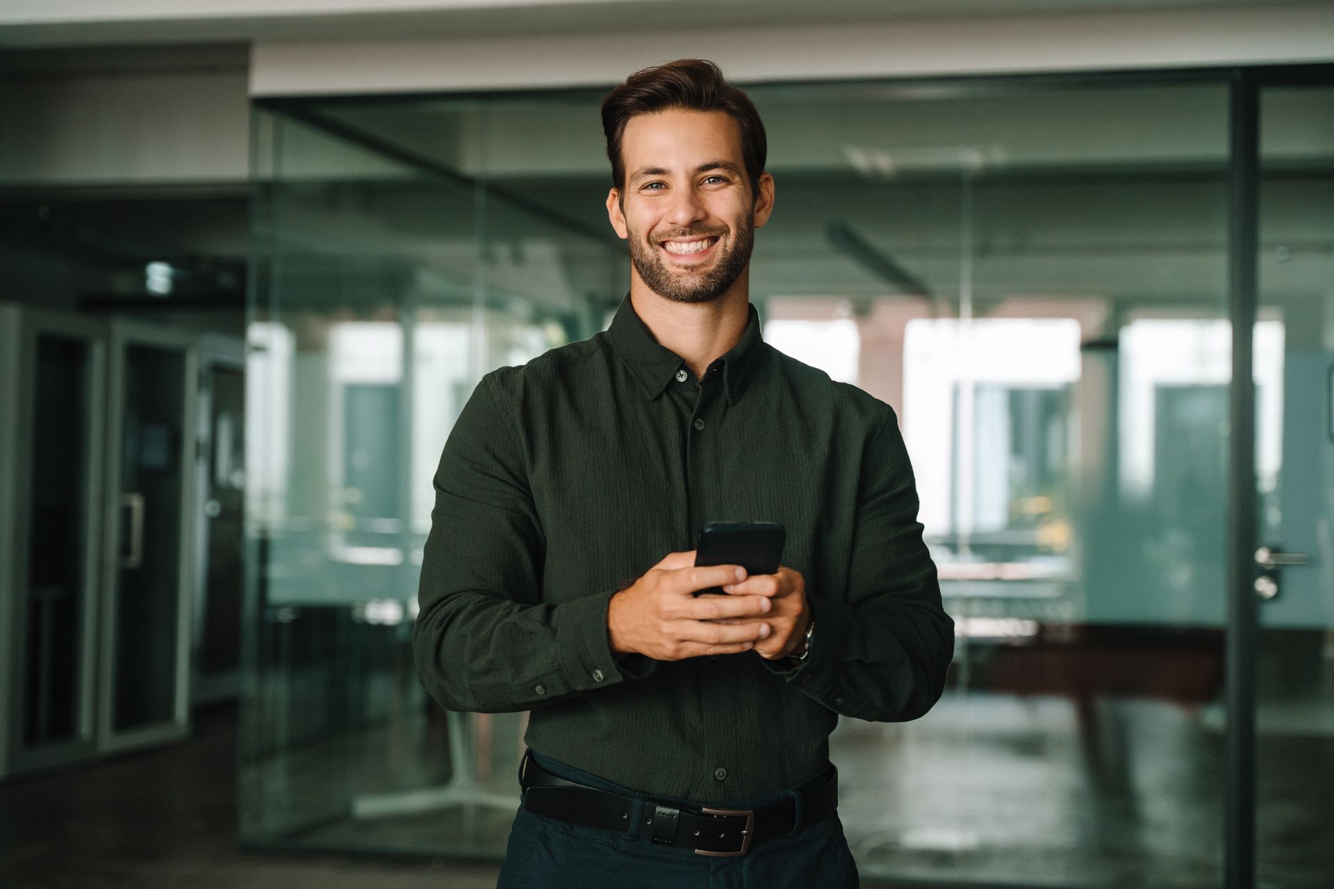 A smiling person wearing a dark green button-down shirt stands in a modern office, holding a smartphone.
