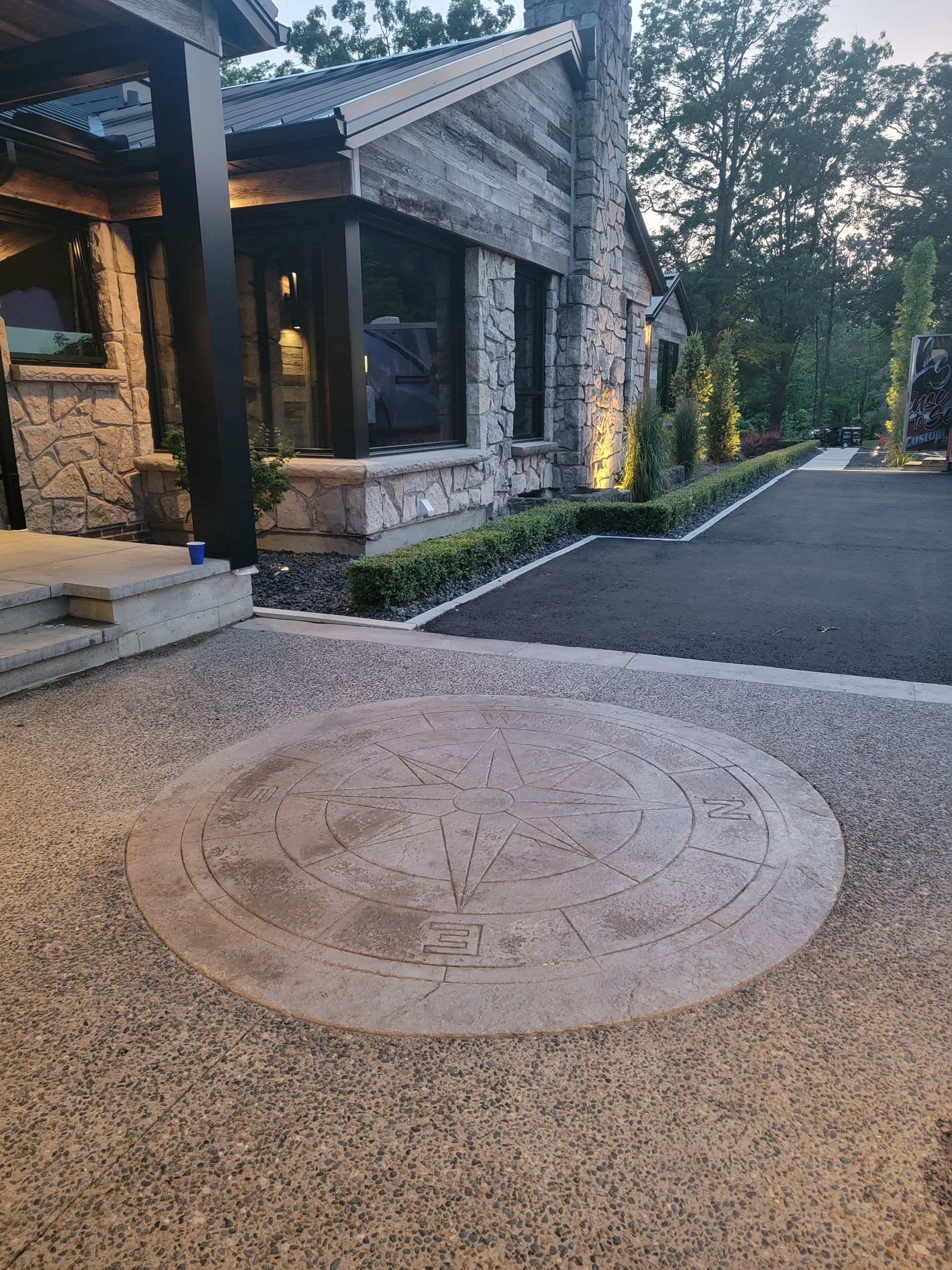 Stone house exterior with a textured circle design on the pavement. Black driveway and green bushes.
