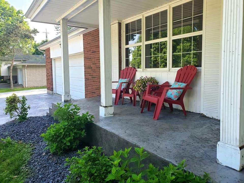 Red chairs on a covered porch with a brick wall and garage, surrounded by landscaping.