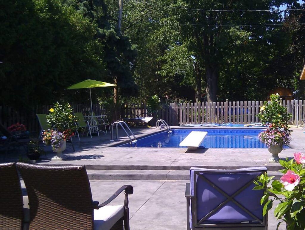 Backyard pool with diving board, patio seating, and fence, surrounded by trees and flowers.