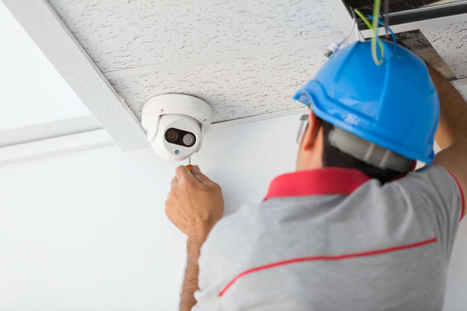 A technician in a blue hard hat installs a security camera on a white ceiling.