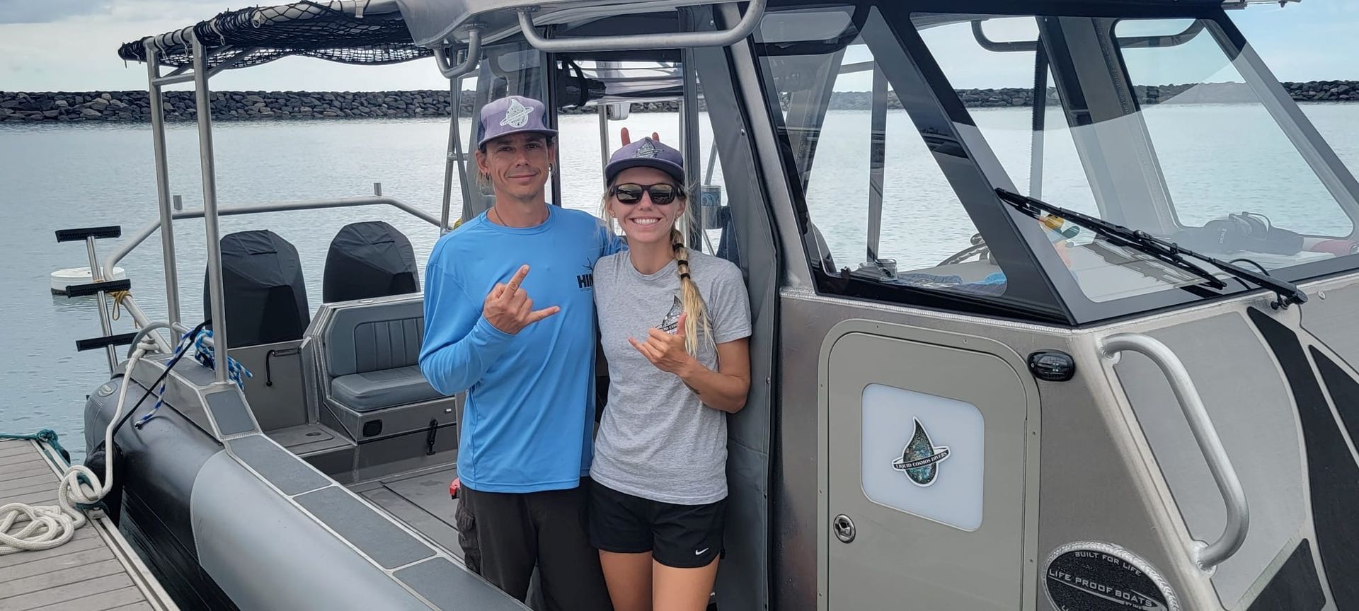 Two people stand beside a boat. They are making hand gestures. They are wearing hats and standing near a body of water.