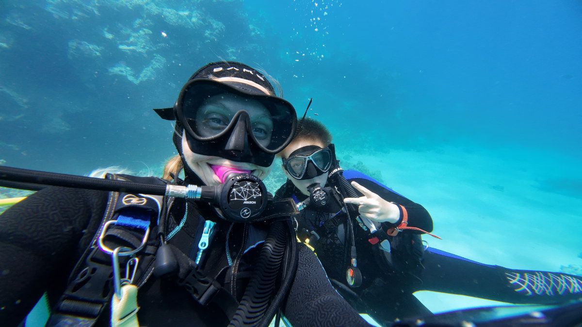 Two people scuba diving, taking a selfie underwater. One smiles, the other gives a peace sign, blue water, and sandy seabed.
