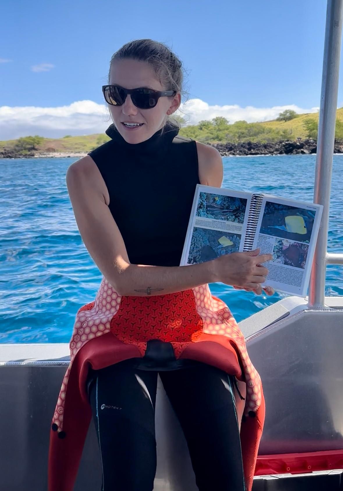 A woman in scuba gear is sitting on a boat holding a book.