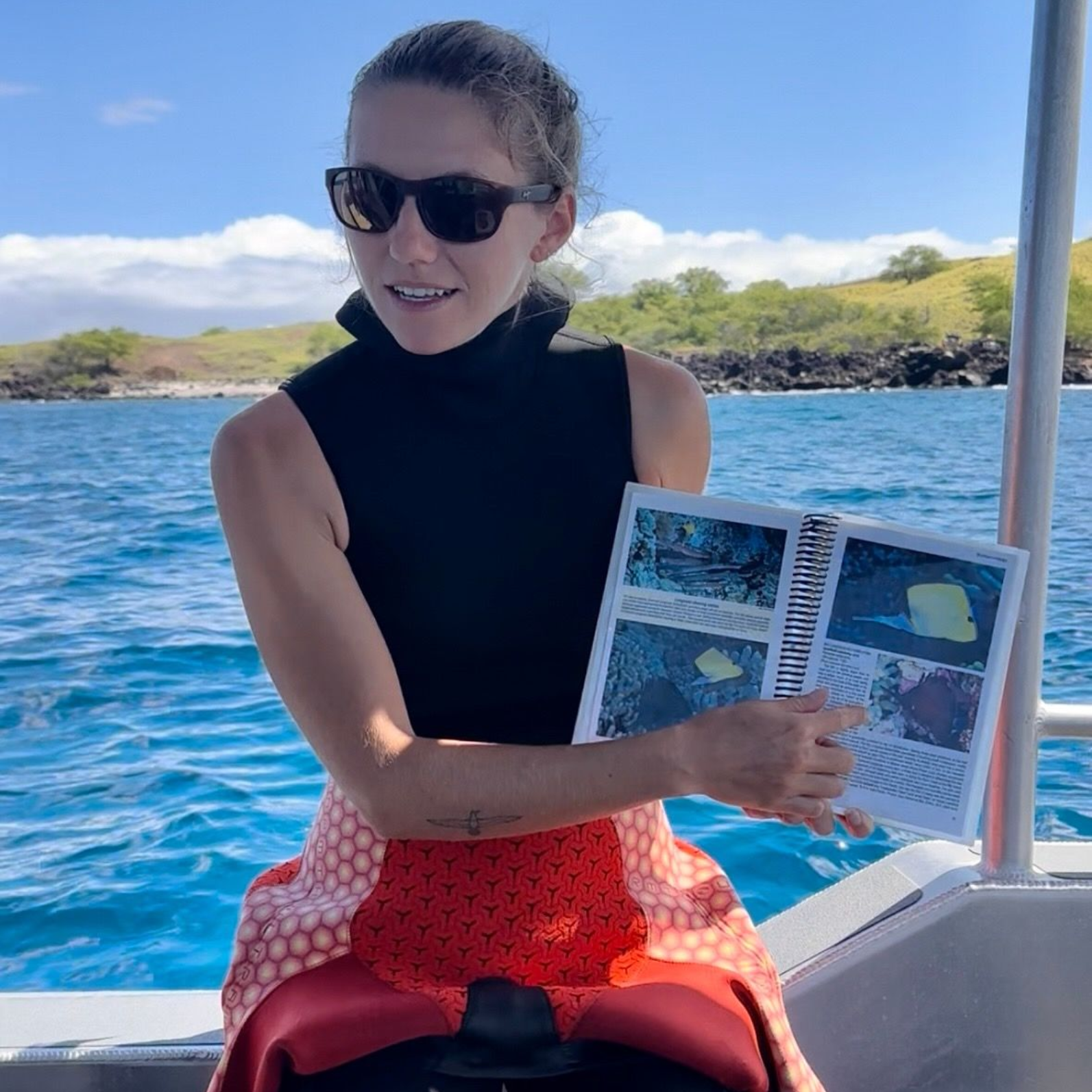Woman on boat, holding educational booklet, pointing. Wears sunglasses, black top, and red wetsuit. Ocean background.