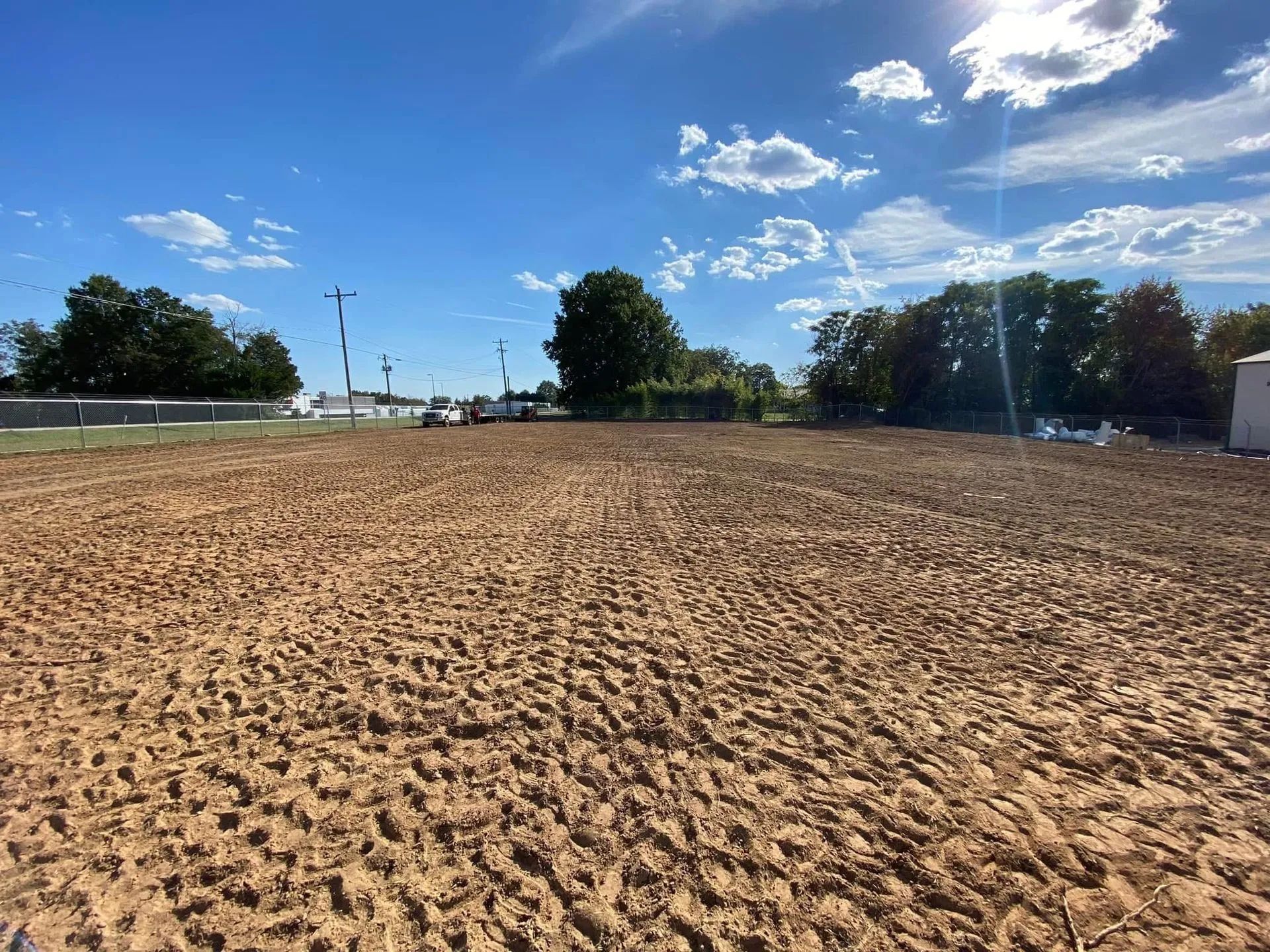 Brown, tilled field under a blue sky with trees and a few structures in the background.