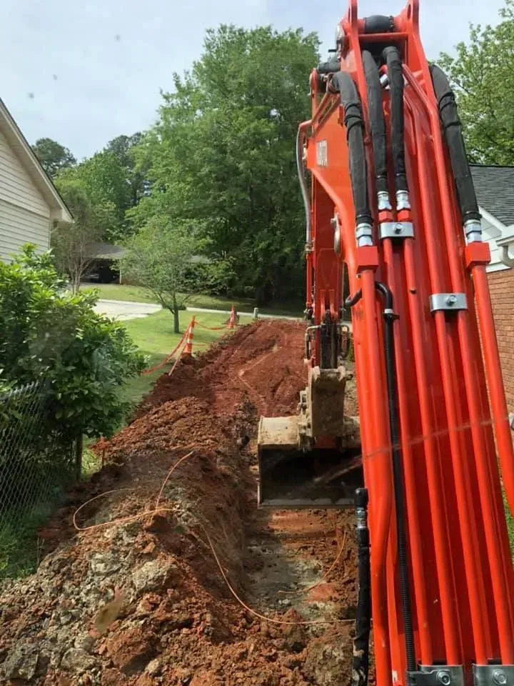 An orange excavator is digging a hole in the dirt in front of a house.