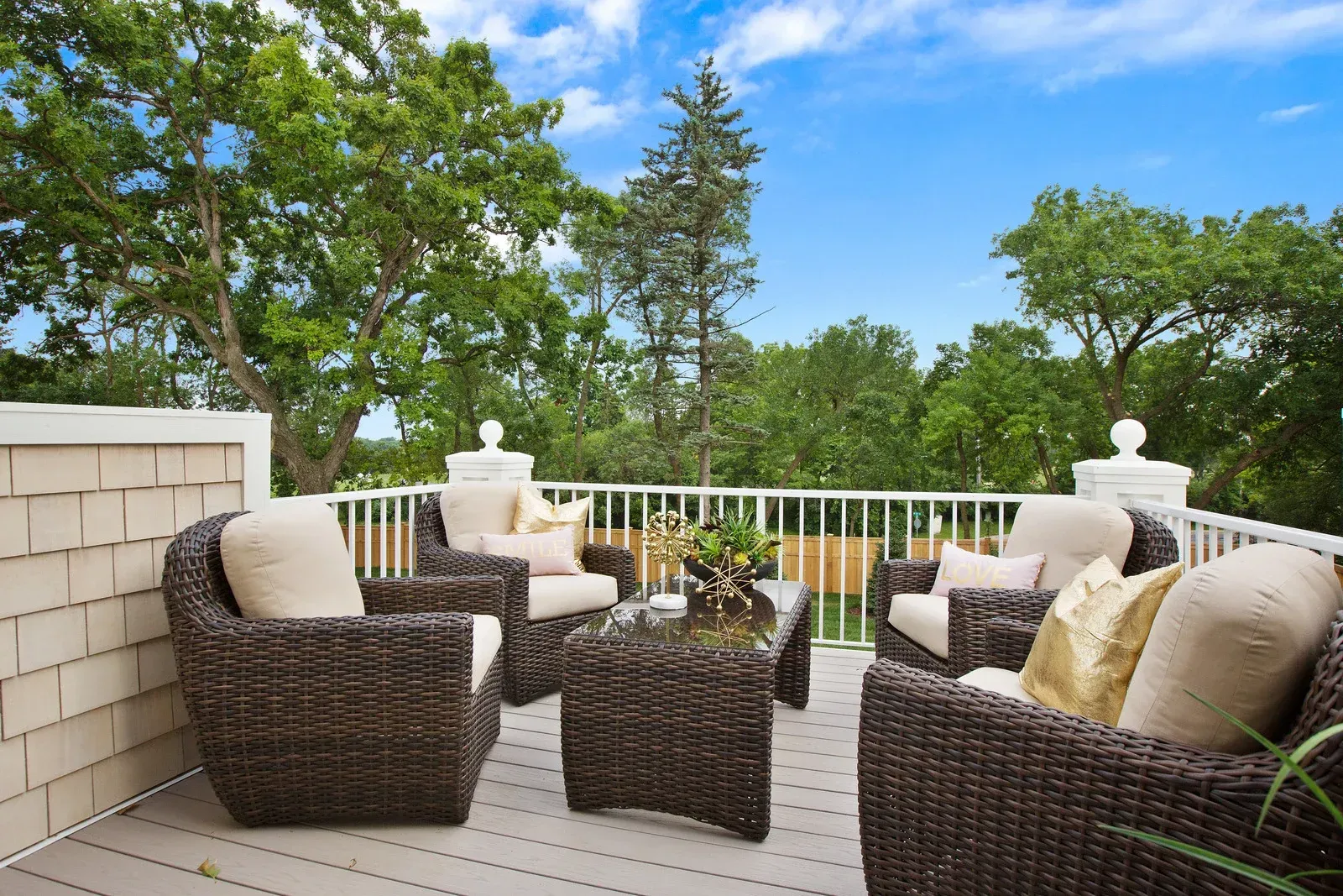 Patio with wicker furniture, white railing, surrounded by trees, under blue sky.