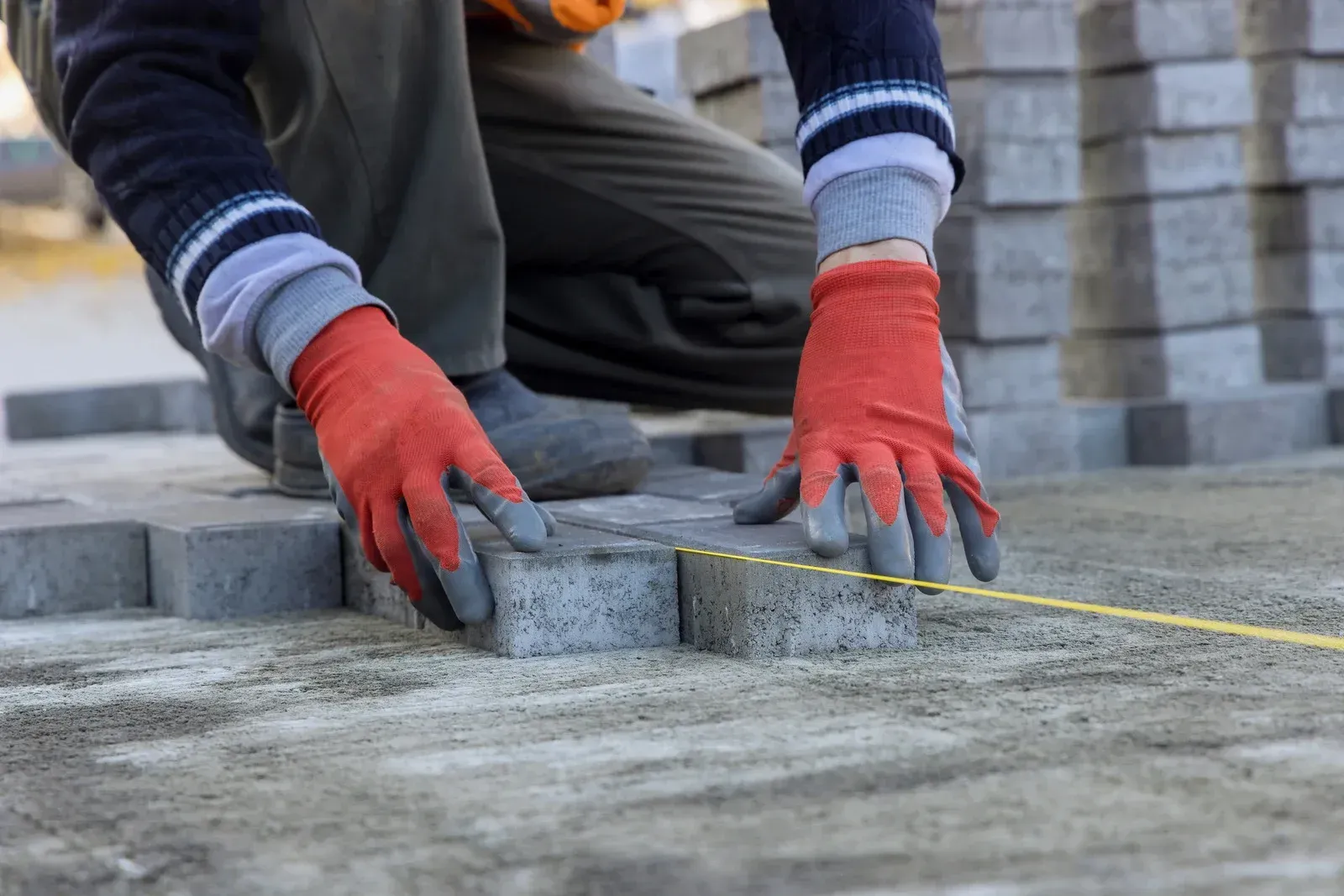Construction worker in orange gloves laying gray paving stones on a cement base.