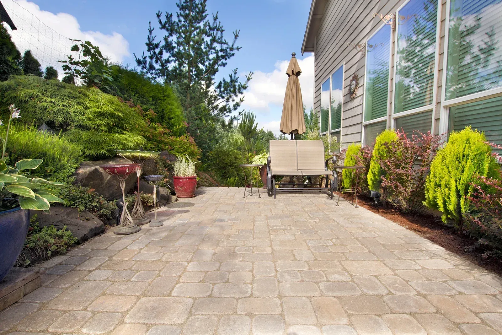 Patio with brick pavers, beige umbrella over a bench, surrounded by lush greenery.
