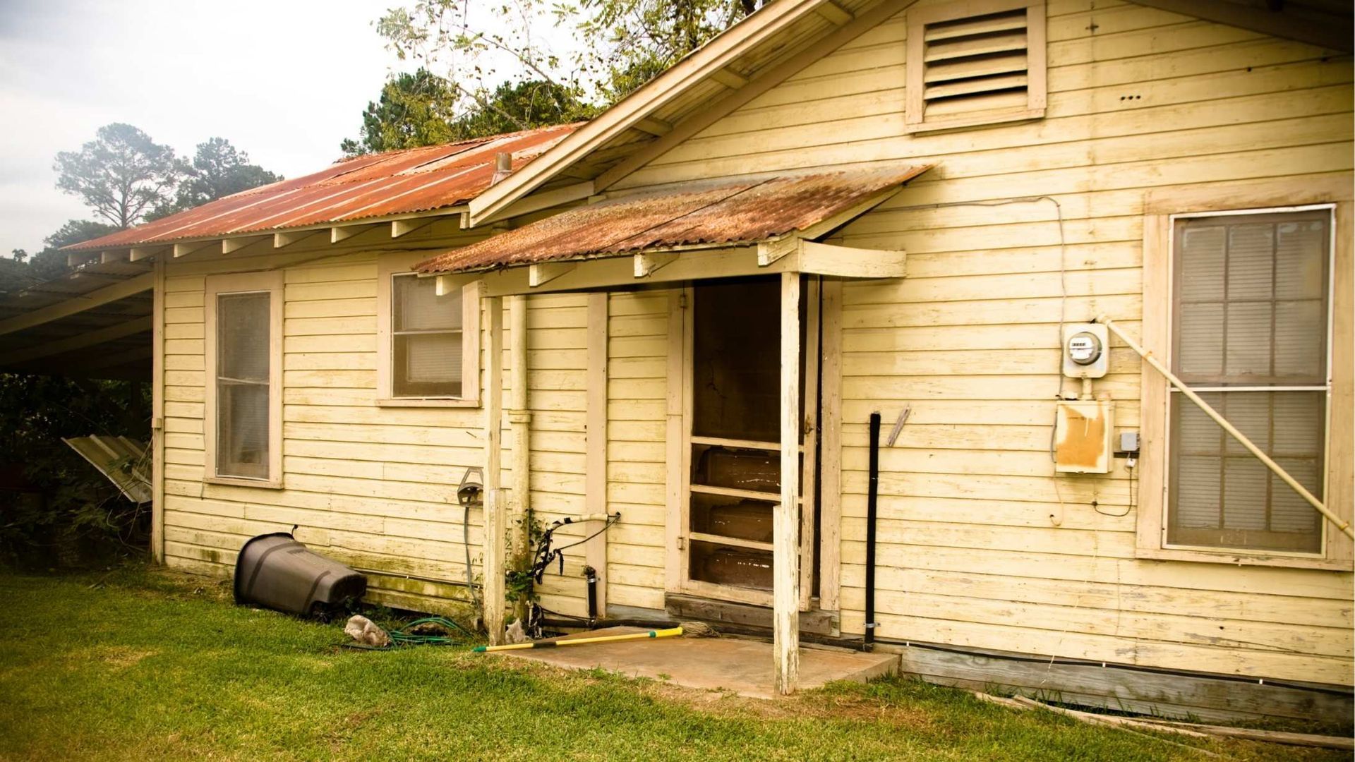 Yellow, weathered house with rusty roof, porch, and screen door; grassy yard.