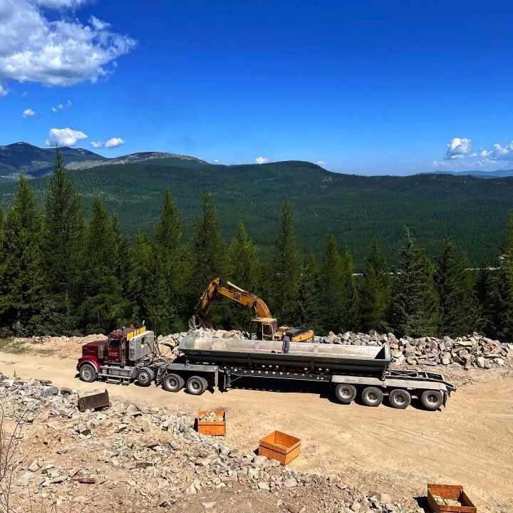 A dump truck is driving down a dirt road with mountains in the background.