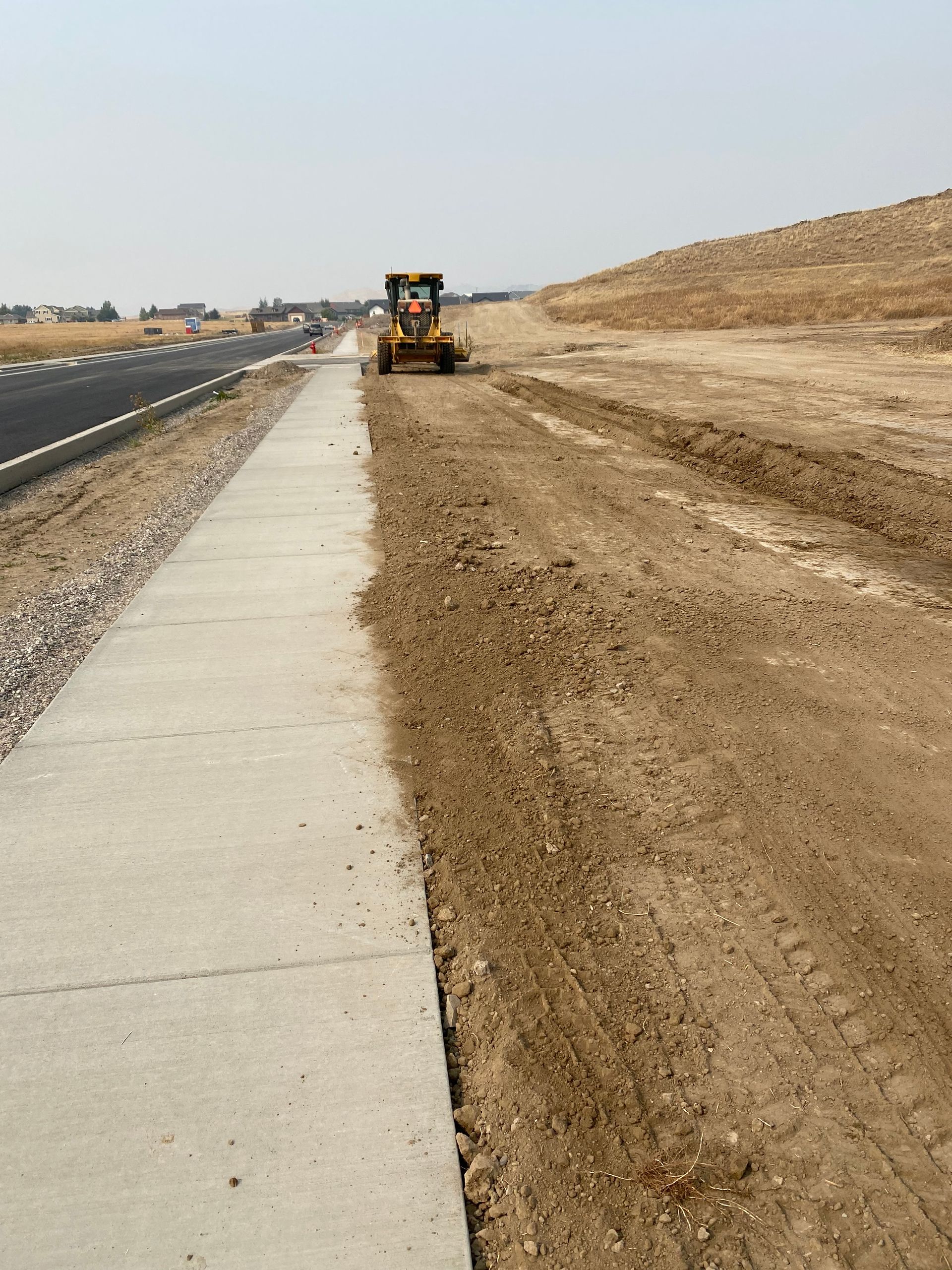 A bulldozer is driving down a dirt road next to a sidewalk.
