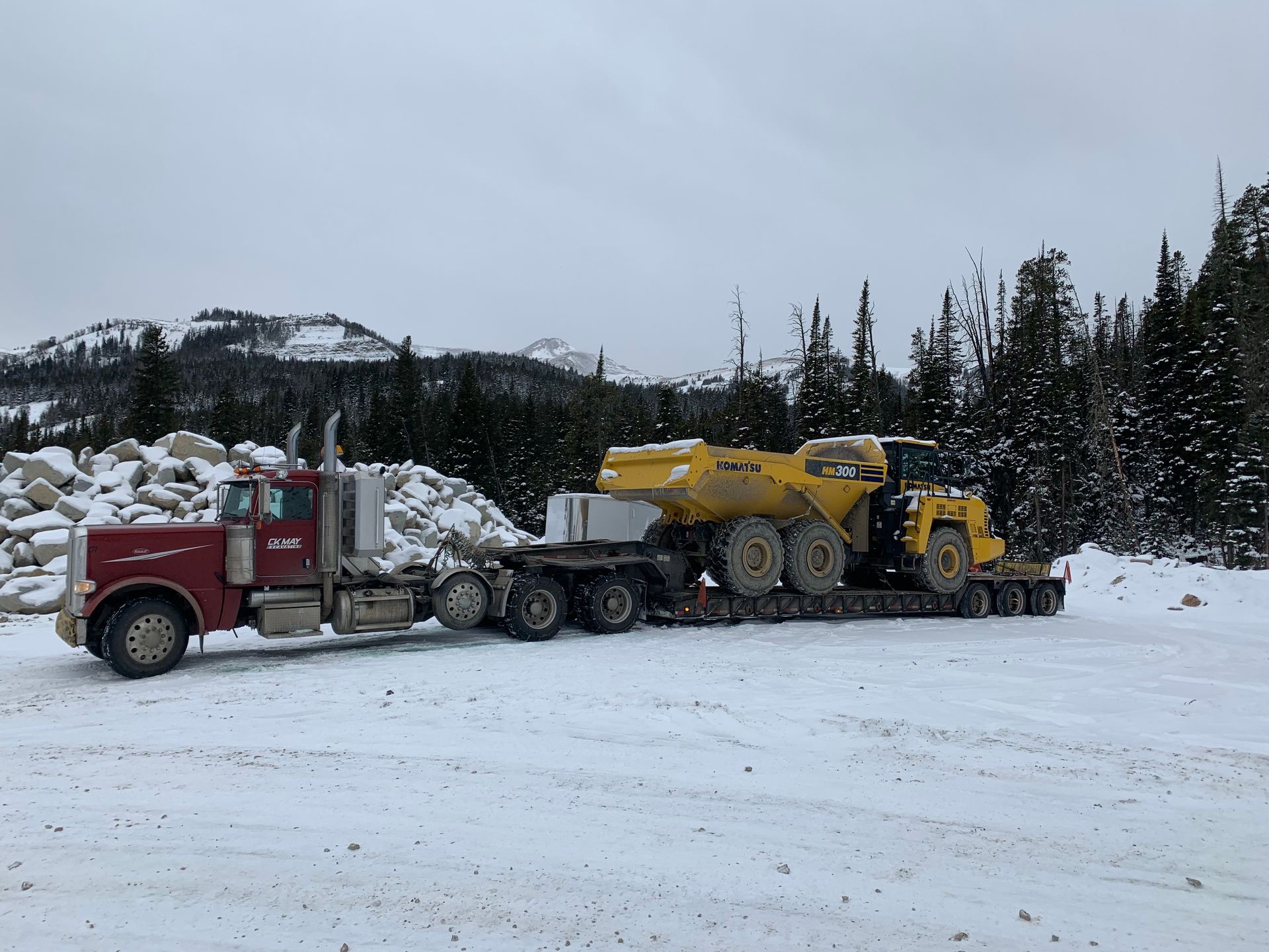 A red semi truck is carrying a yellow dump truck on a snowy road.