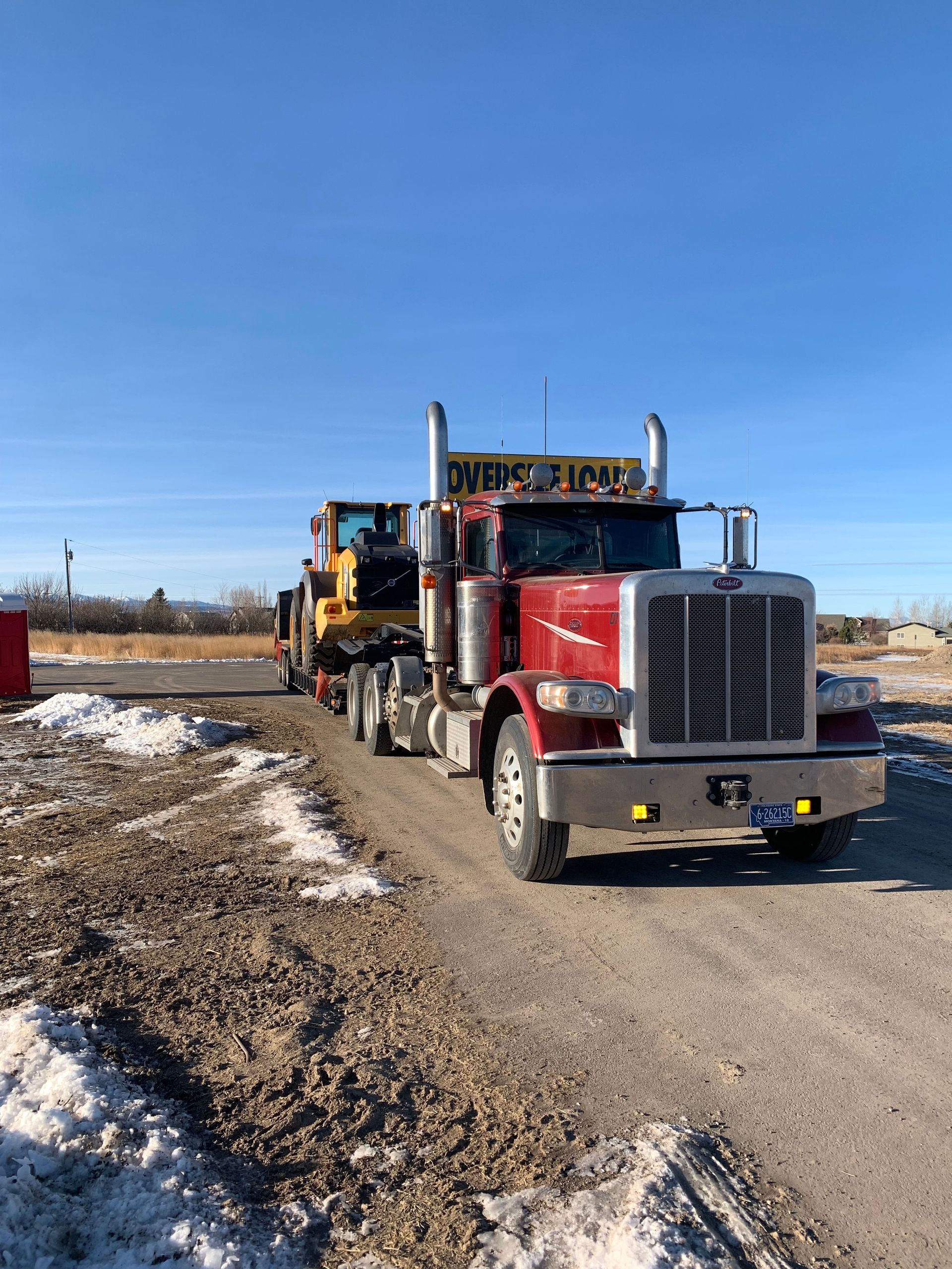 A red semi truck is driving down a dirt road carrying a bulldozer.