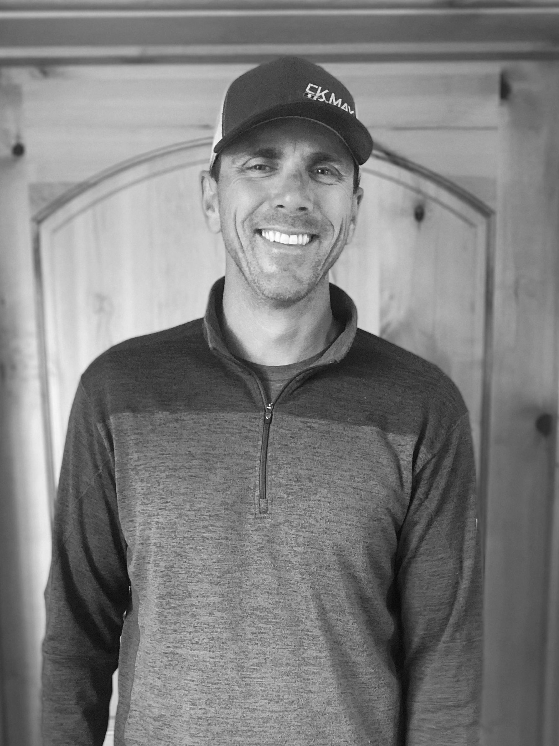 A man wearing a hat and a sweater is smiling in a black and white photo.