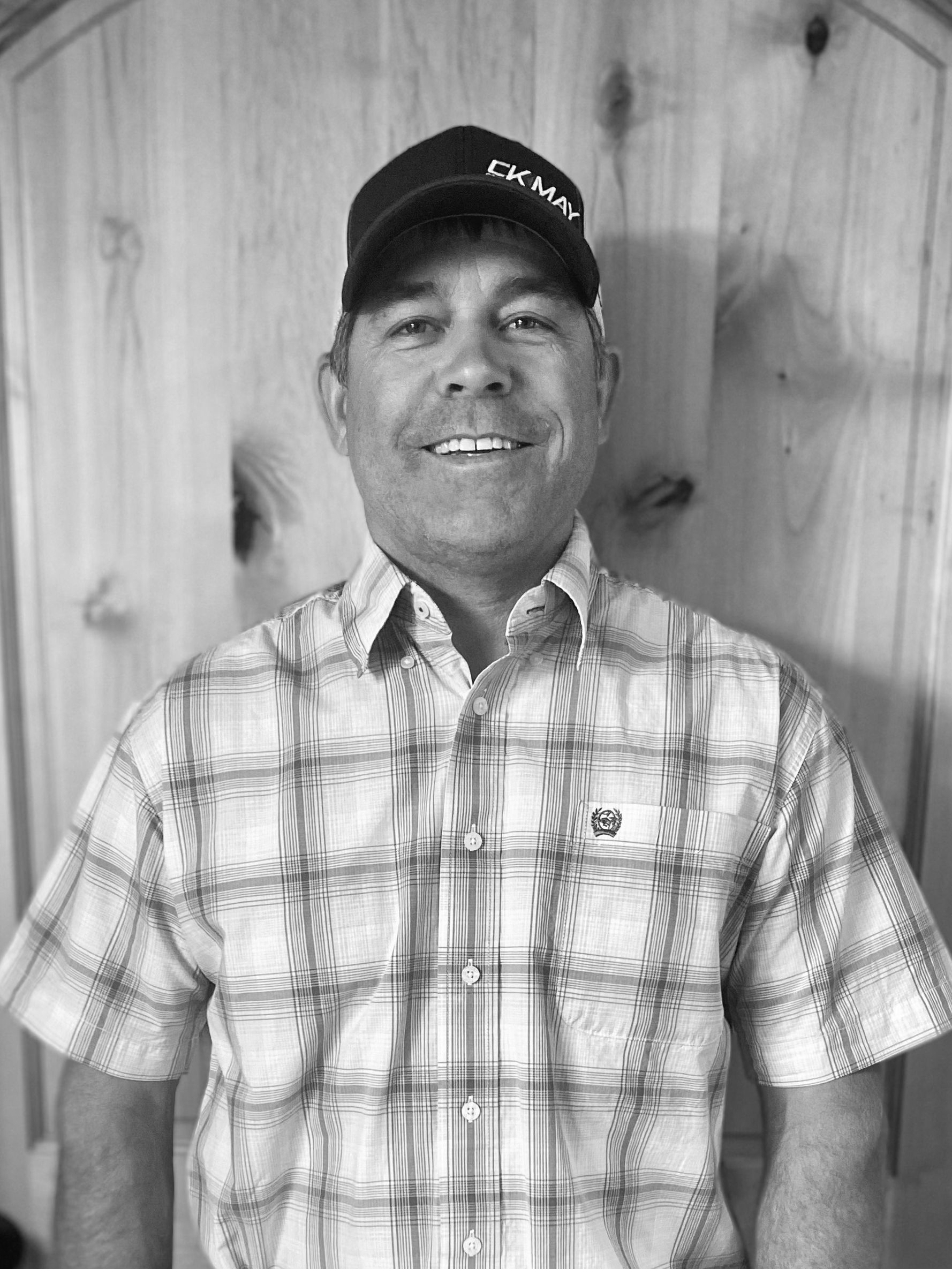 A man wearing a plaid shirt and a hat is smiling in a black and white photo.