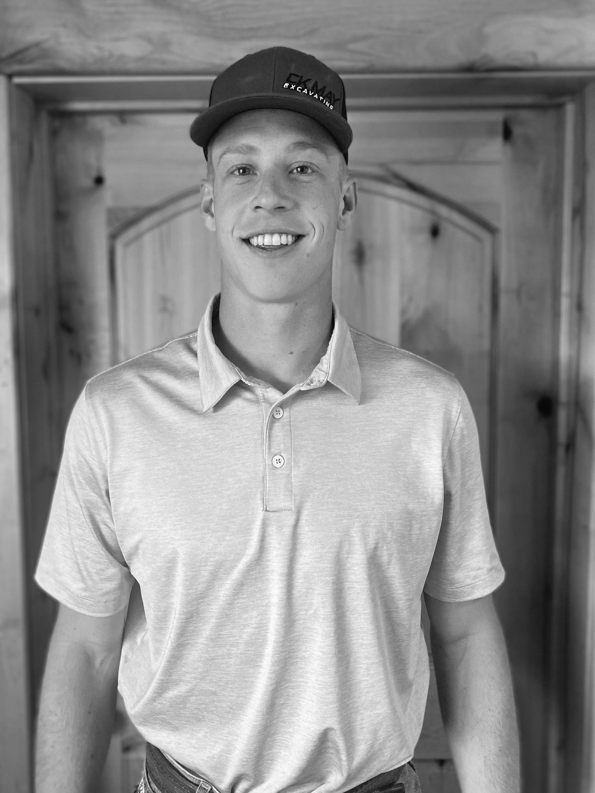 A young man wearing a hat and a polo shirt is standing in front of a wooden door.