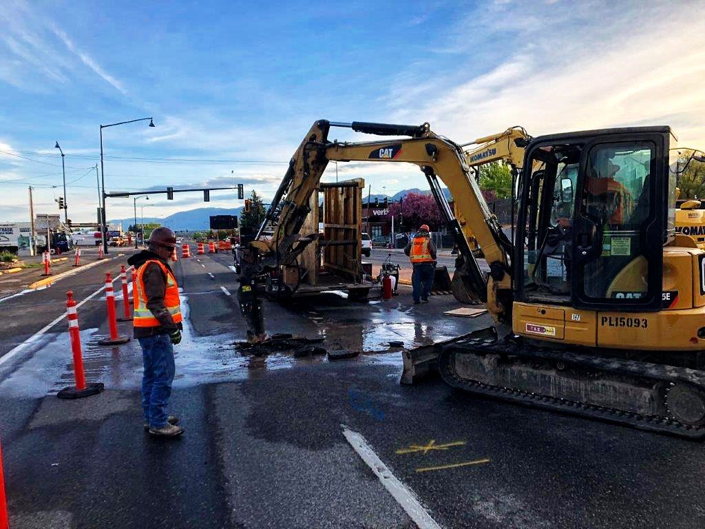 A man in an orange vest is standing next to a yellow excavator on the side of a road.