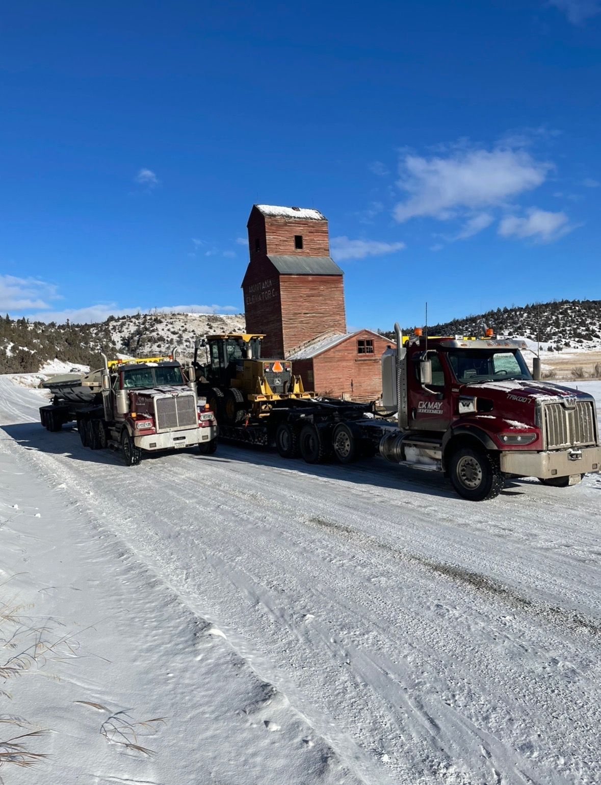 Two trucks are parked on a snowy road in front of a brick building.