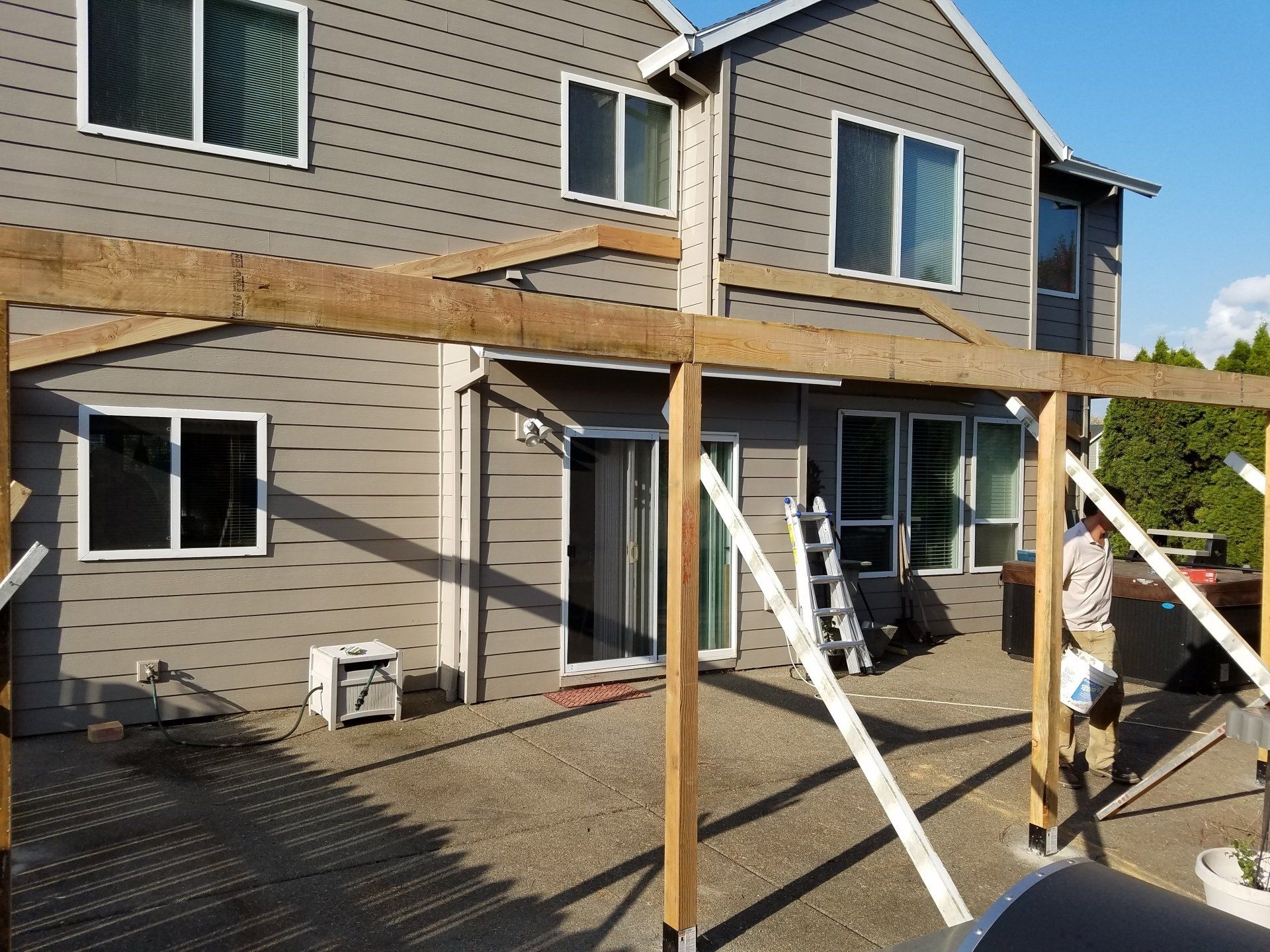 Pergola under construction next to a two-story gray house with windows. A person works on the structure.
