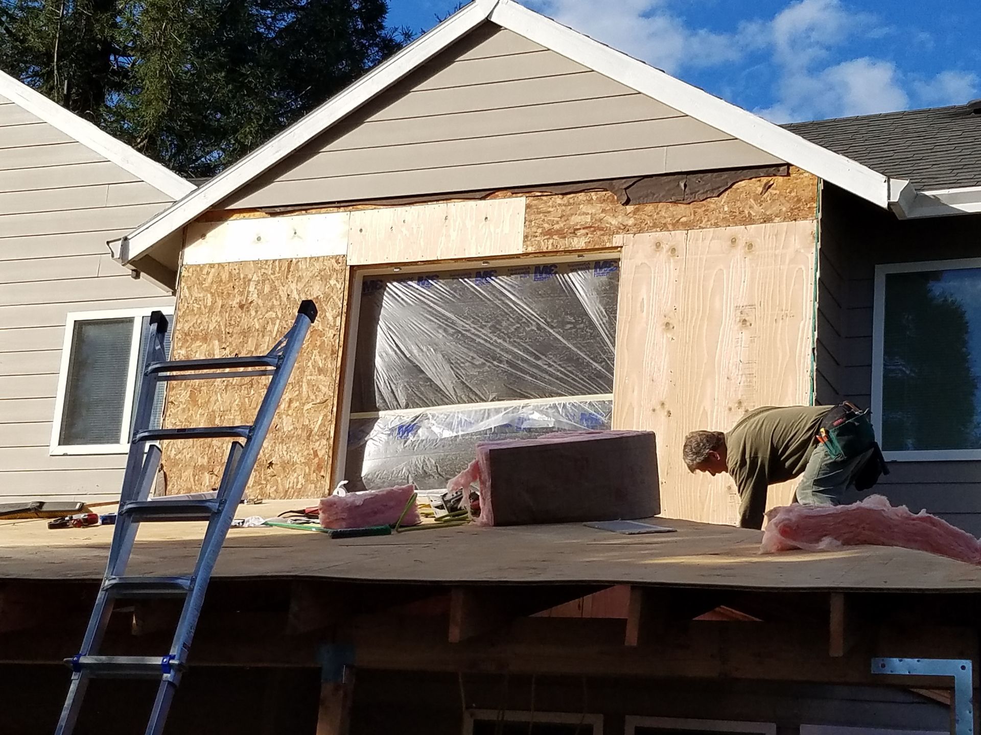 Construction worker on a roof adding plywood to a house, window area exposed with insulation.