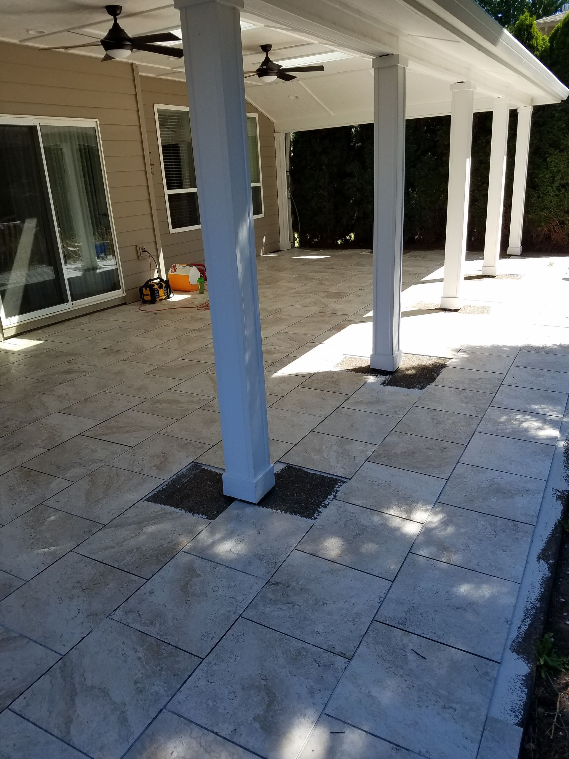 A patio with white columns and pavers under a covered area, next to a building and greenery.