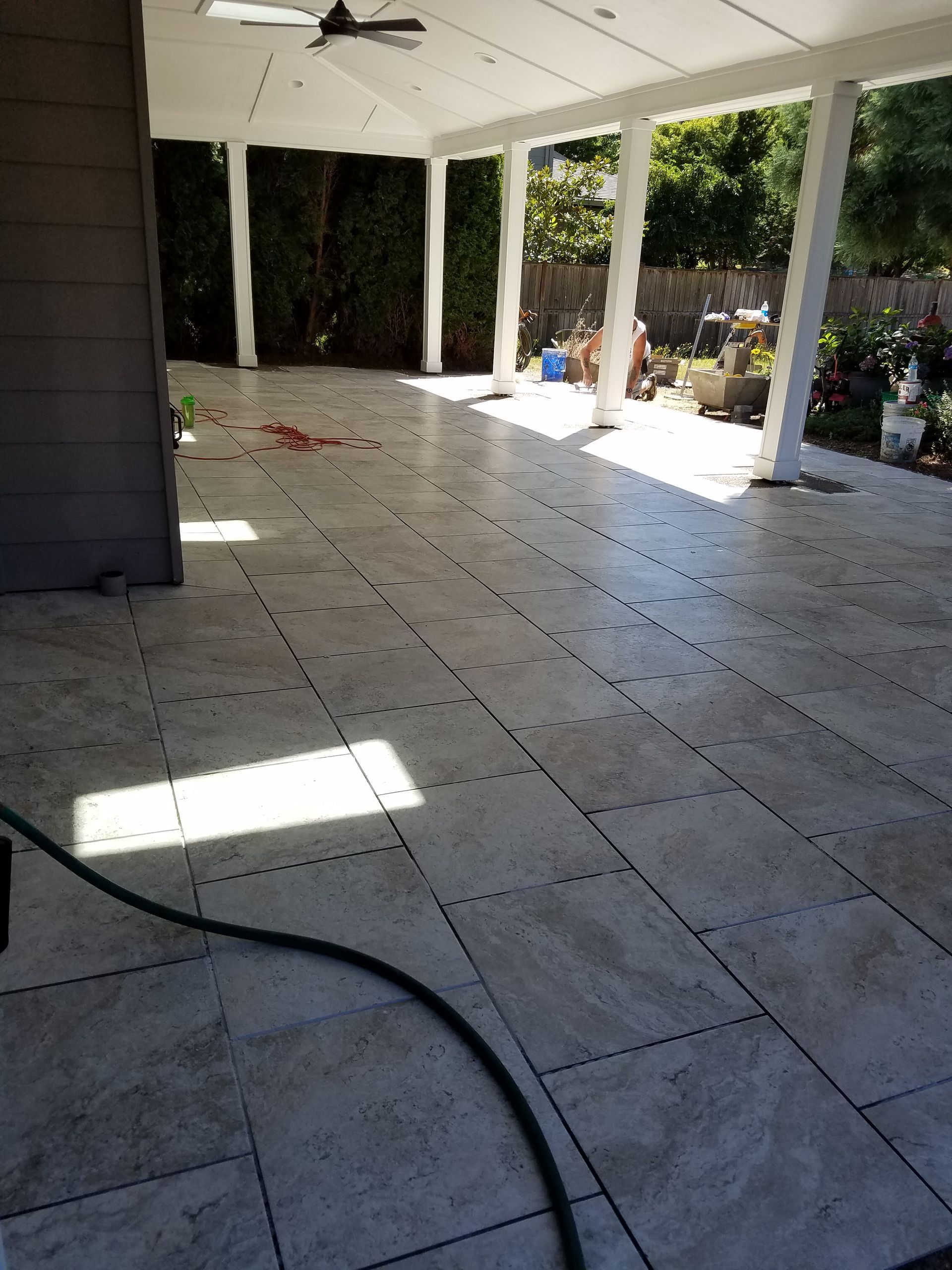 Covered outdoor patio with light gray tile flooring, white pillars, and green foliage in the background.