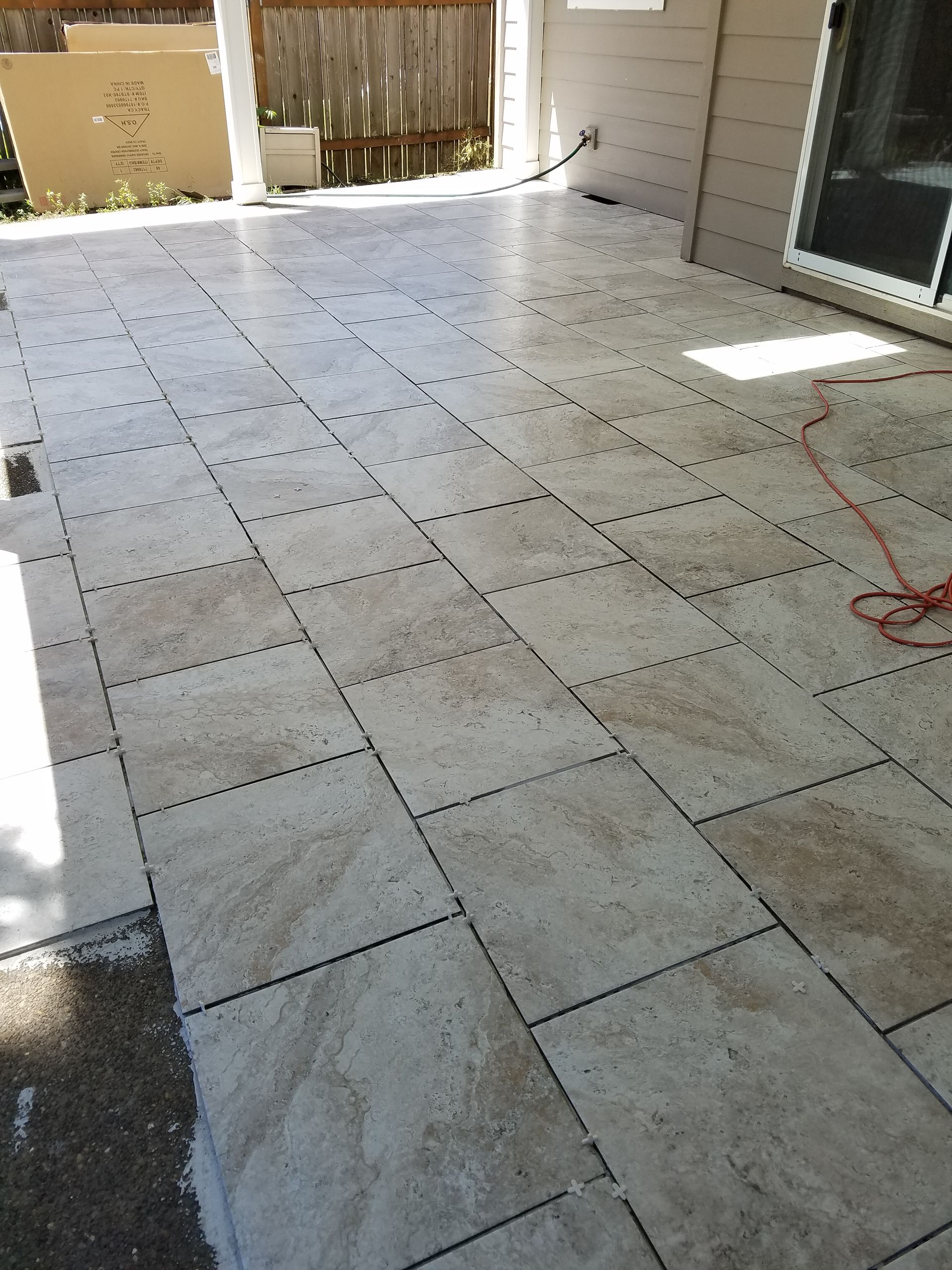 Patio with tan rectangular tiles, partially covered, near a sliding glass door and fence.