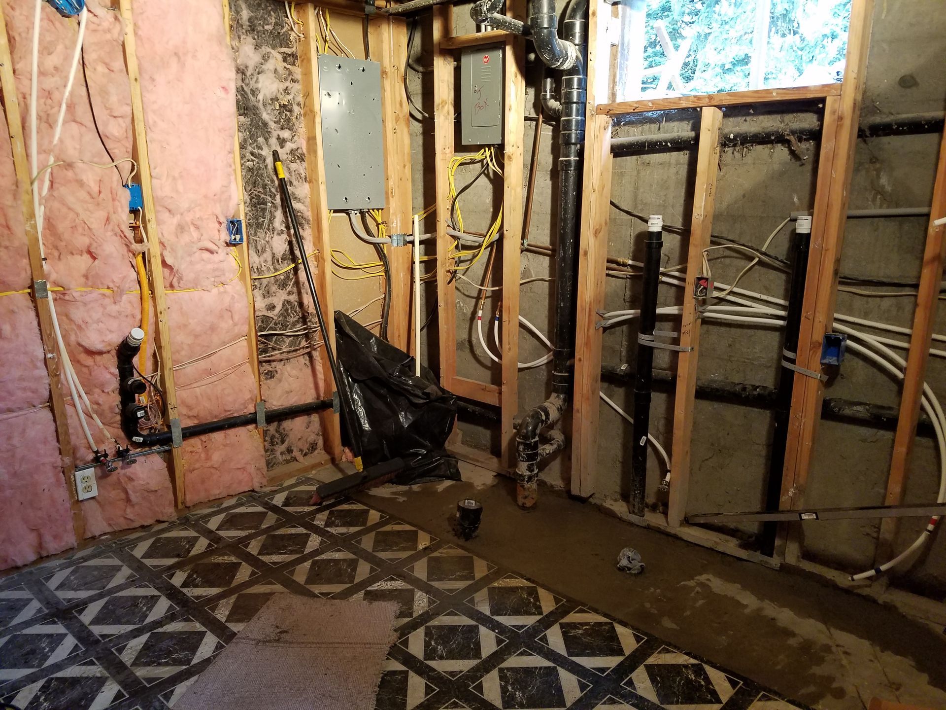 Unfinished room renovation, exposed studs, insulation, electrical wiring, and plumbing. Black pipes, flooring tiles, and a trash bag are present.