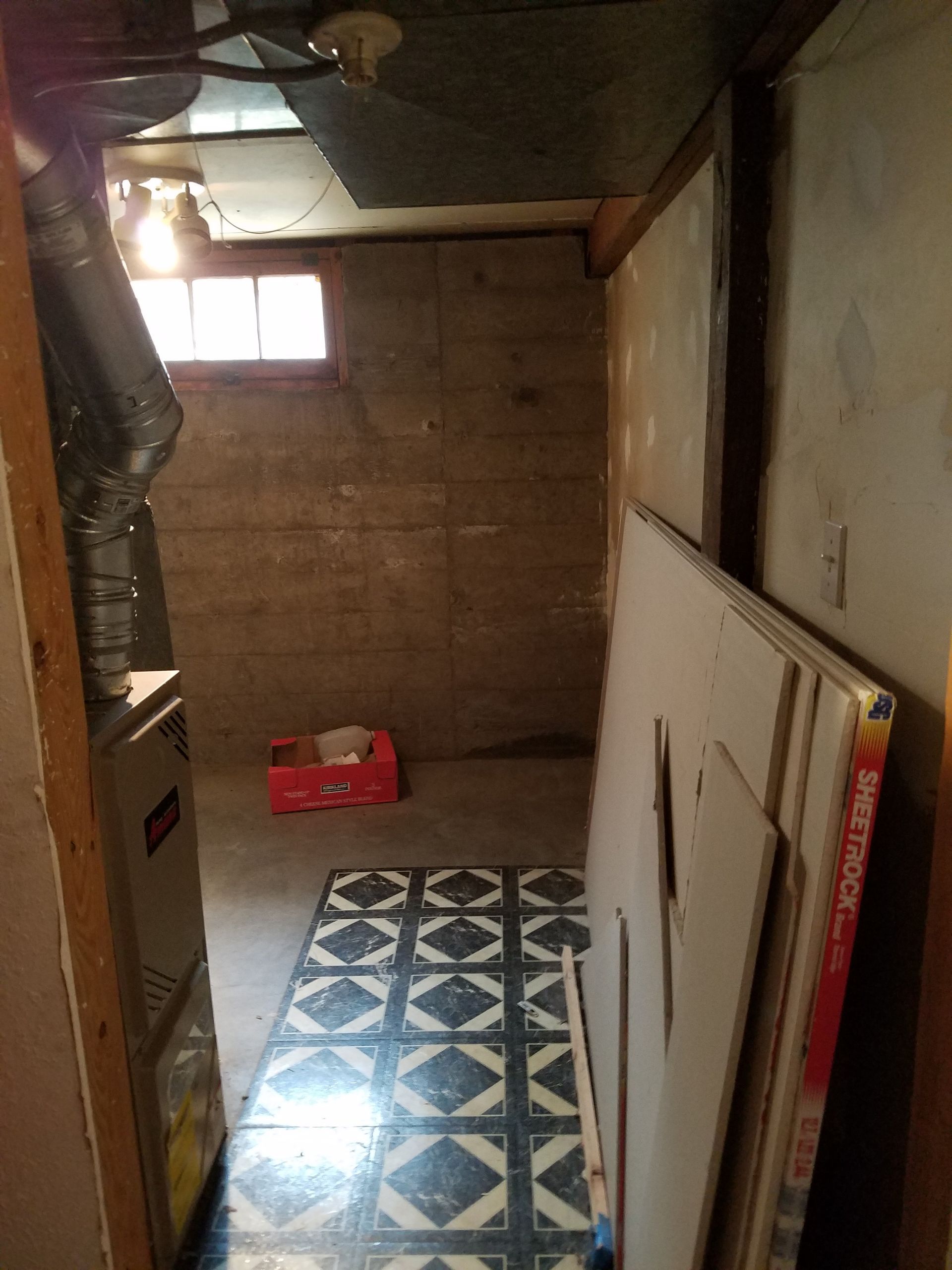 Basement hallway with furnace, small window, and stacked drywall panels. Dark walls, patterned floor.