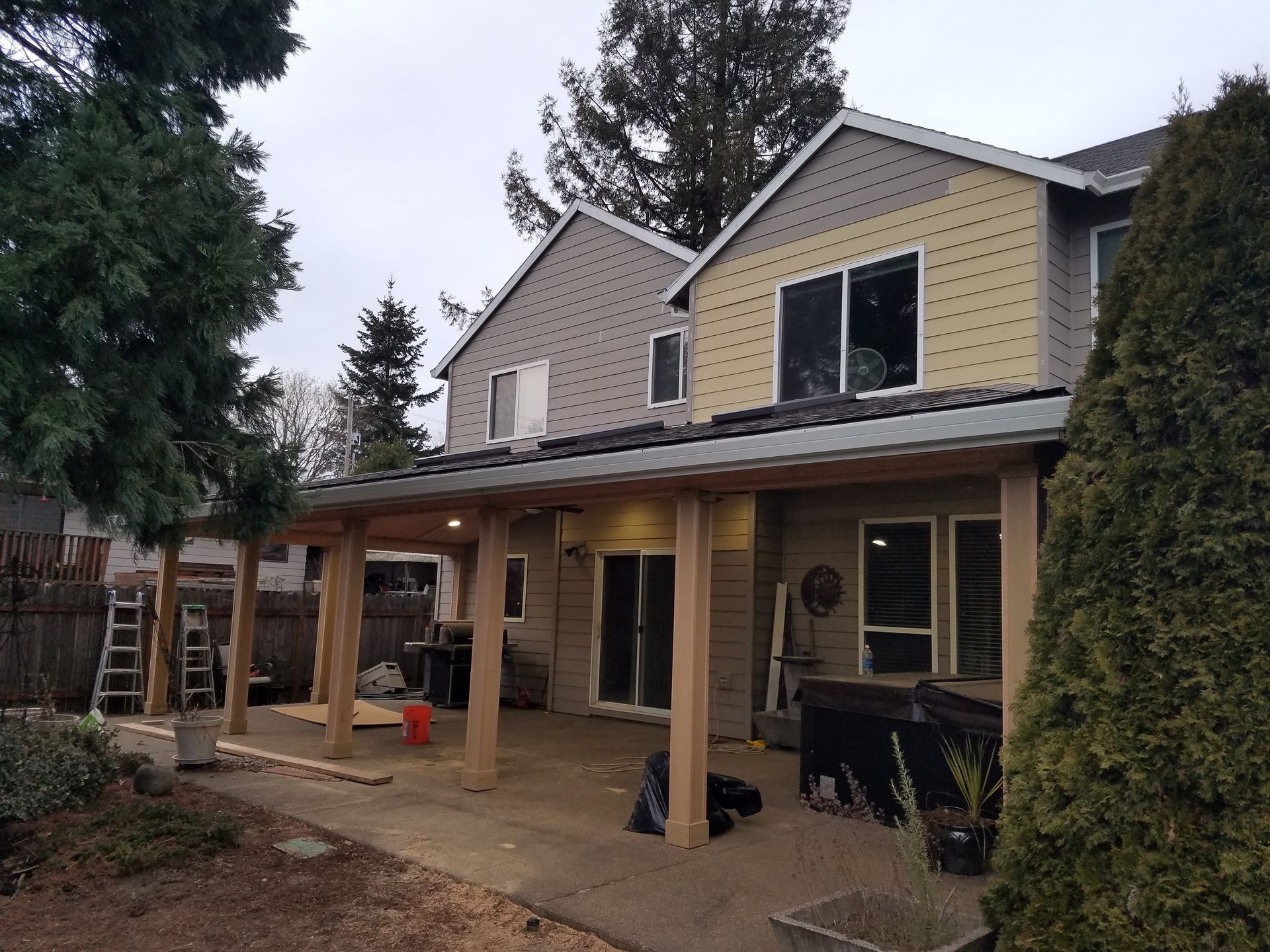 Backyard patio with wooden posts and a partial roof, attached to a two-story house with gray and yellow siding.